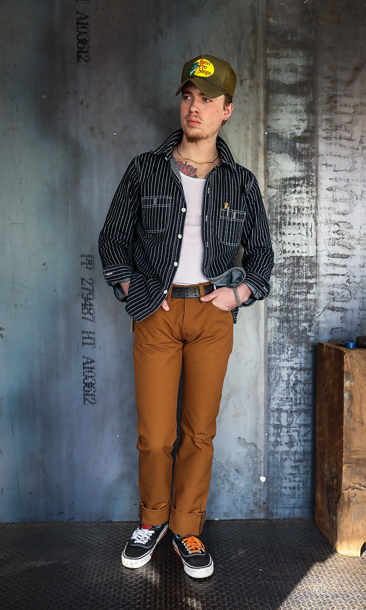Man wearing brown pants, black striped jacket, white tank top, and green cap standing against industrial metal wall