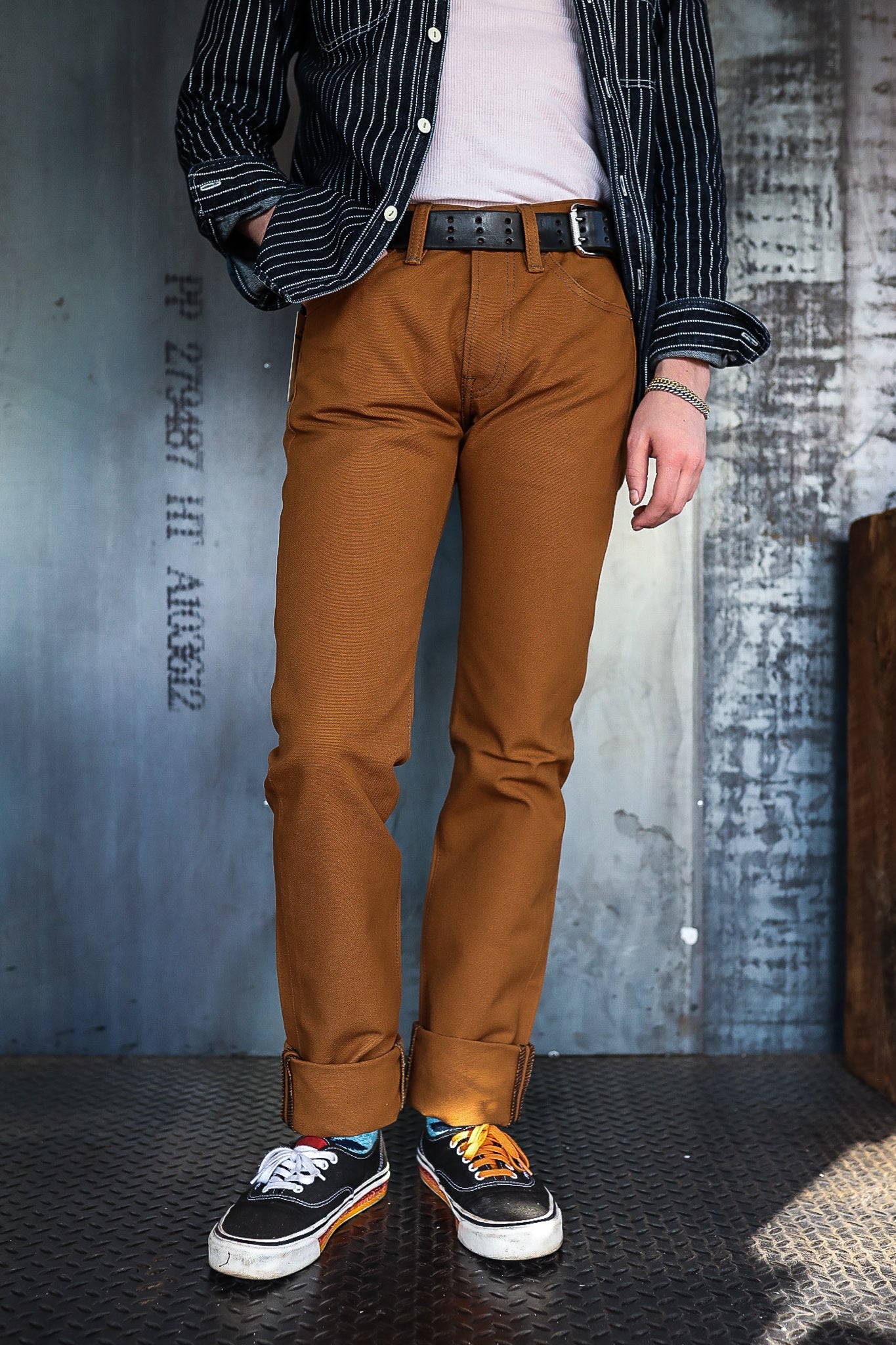 Person wearing brown work pants, black belt, striped black shirt, and mismatched black sneakers on industrial metal floor