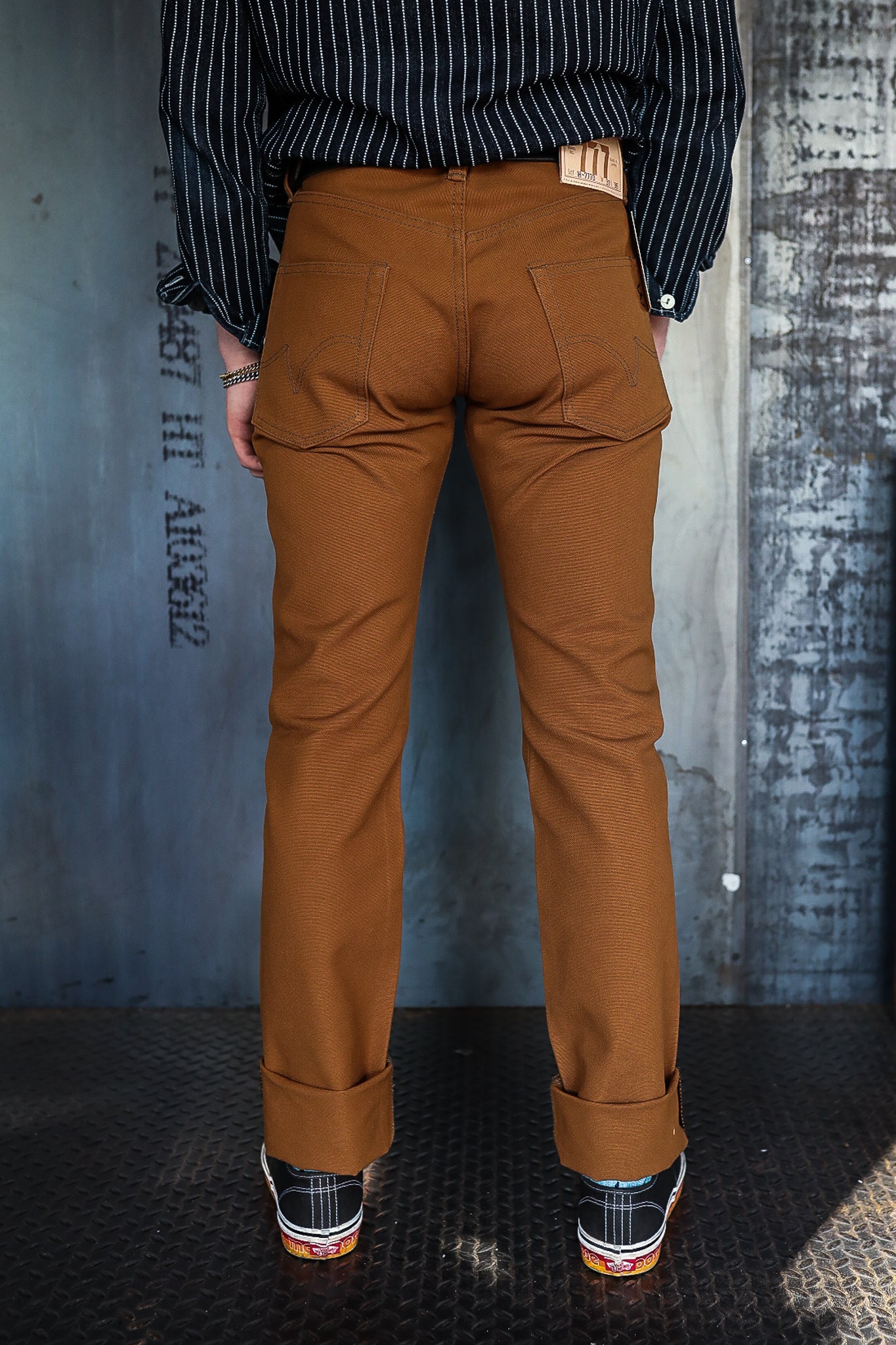 Back view of person wearing brown durable work pants with cuffed hems and black sneakers on industrial floor