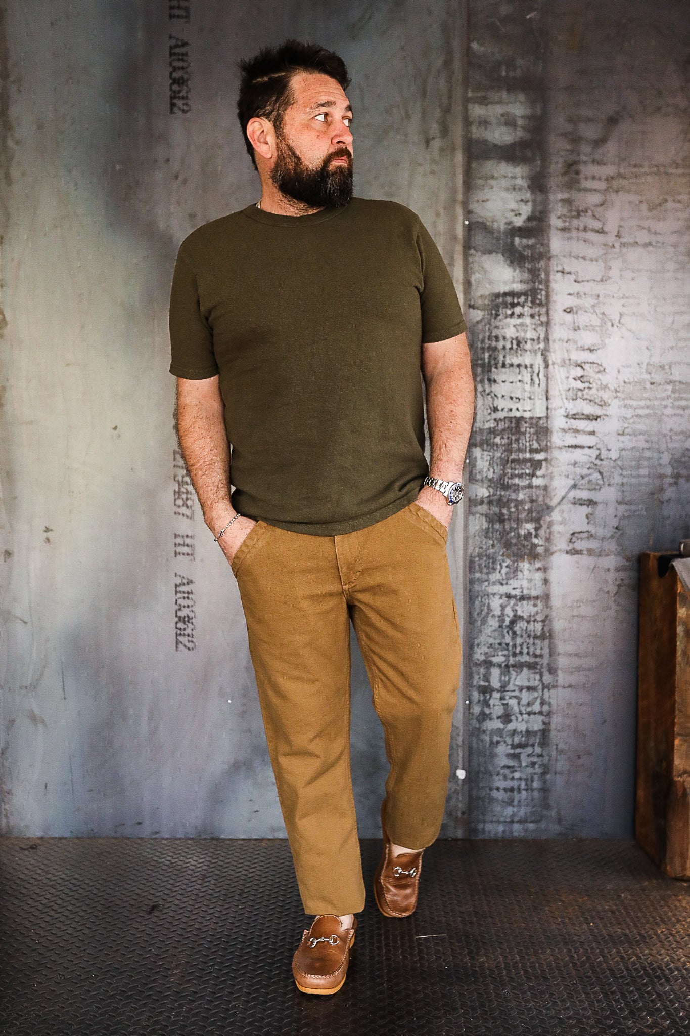 Man in olive green t-shirt and tan pants wearing brown leather loafers standing in industrial setting
