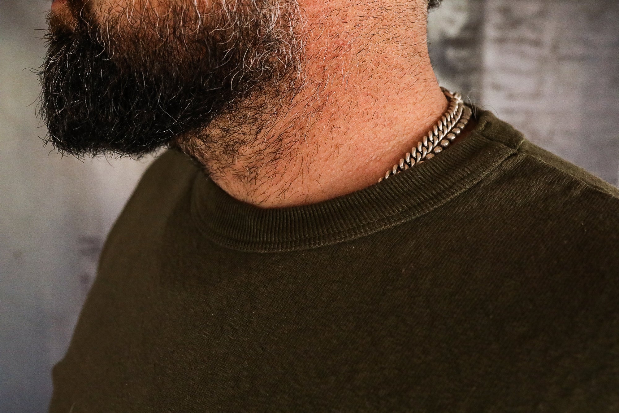 Close-up of a man's neck with a chain necklace, dark beard, and olive green shirt