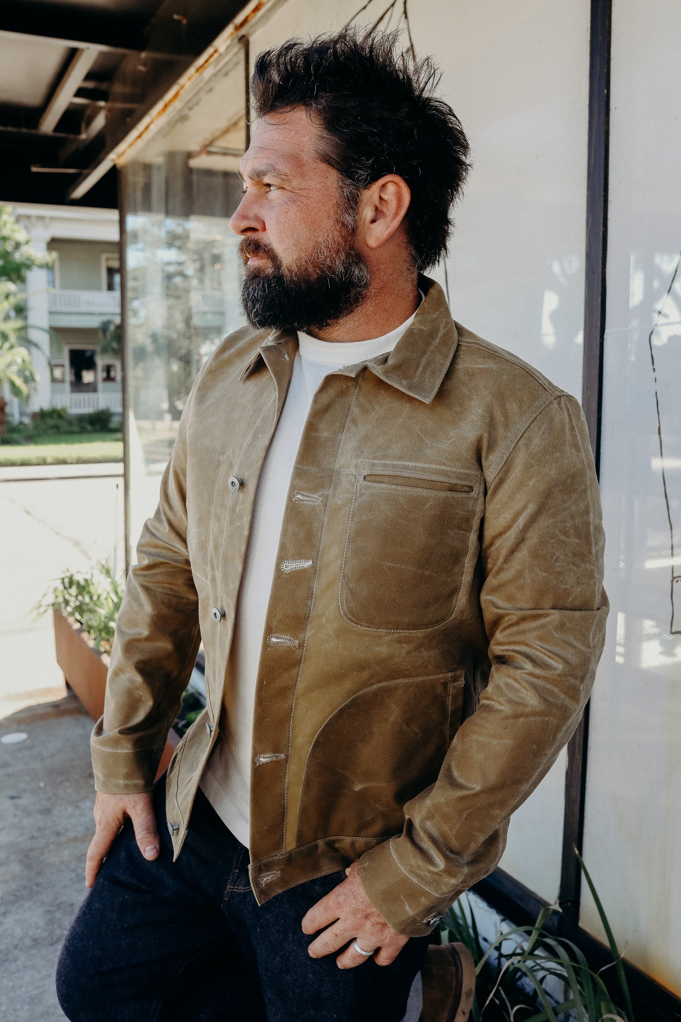 Bearded man wearing a tan leather jacket and white shirt standing outdoors near glass window