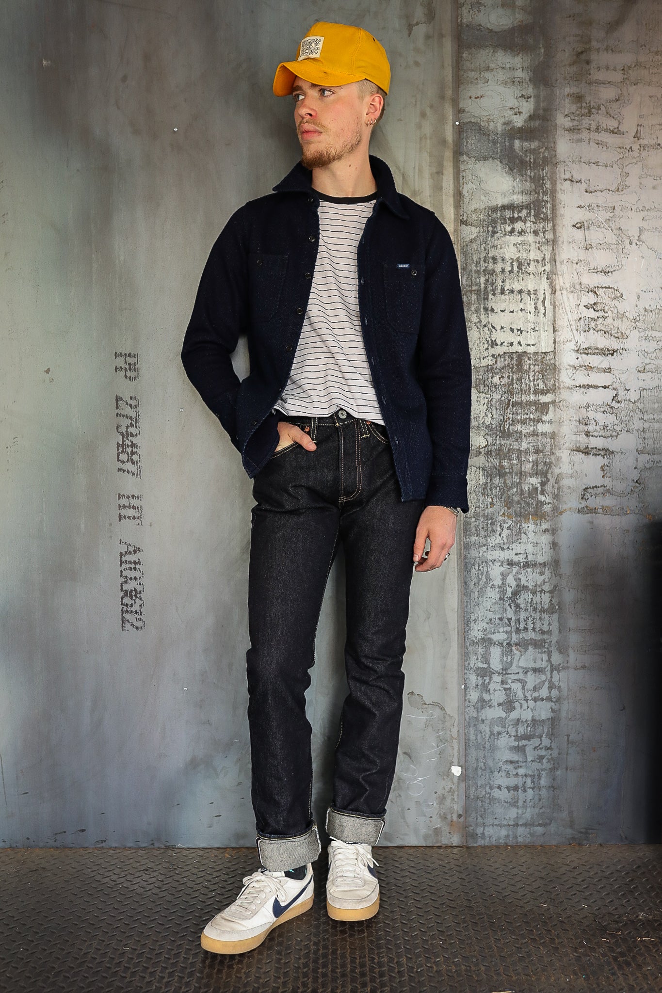 Man wearing a yellow cap, navy jacket, striped shirt, dark rolled-up jeans, and white sneakers in industrial setting