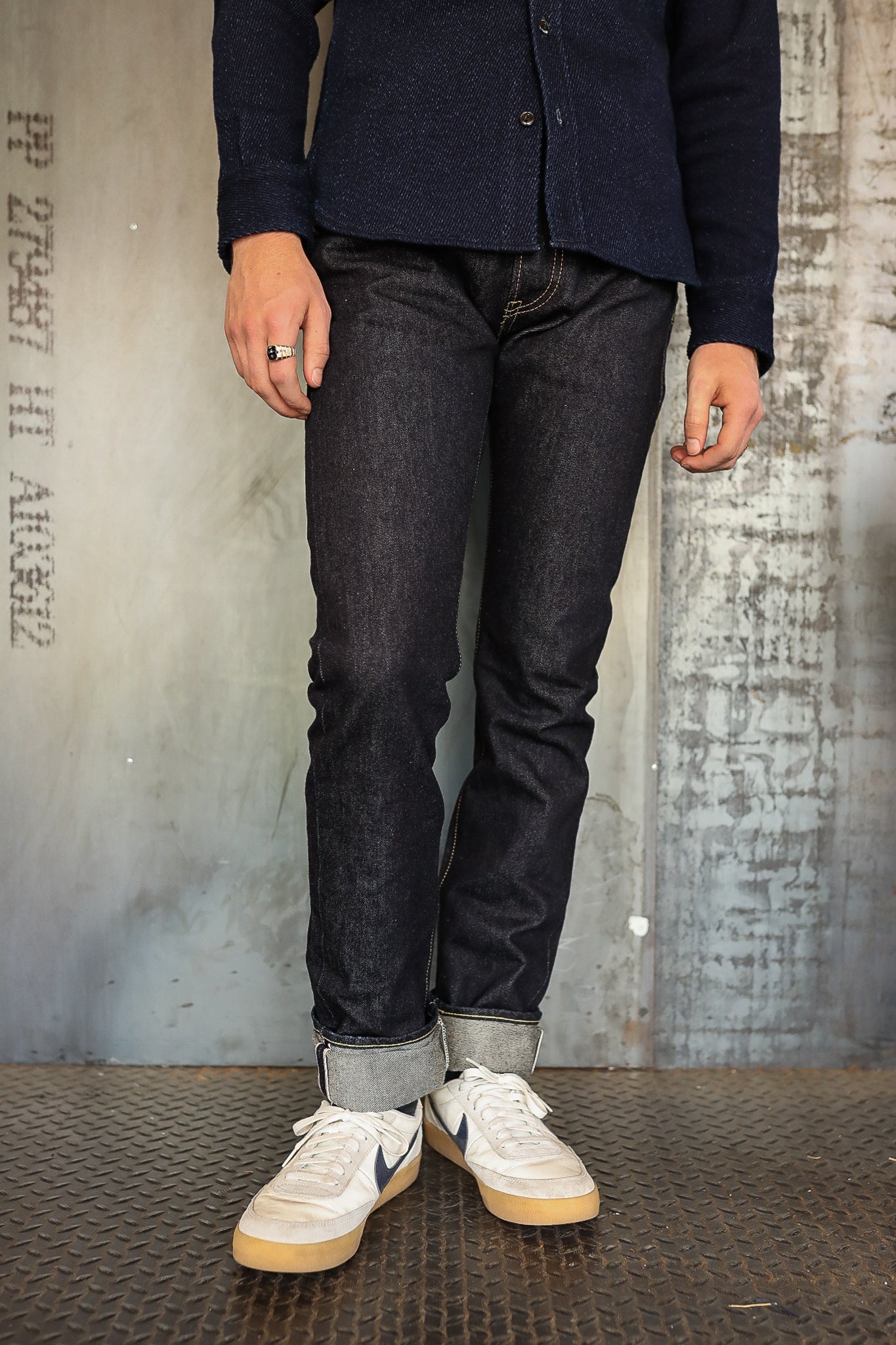 Man wearing dark raw denim jeans with rolled cuffs and white Nike sneakers standing on metal floor