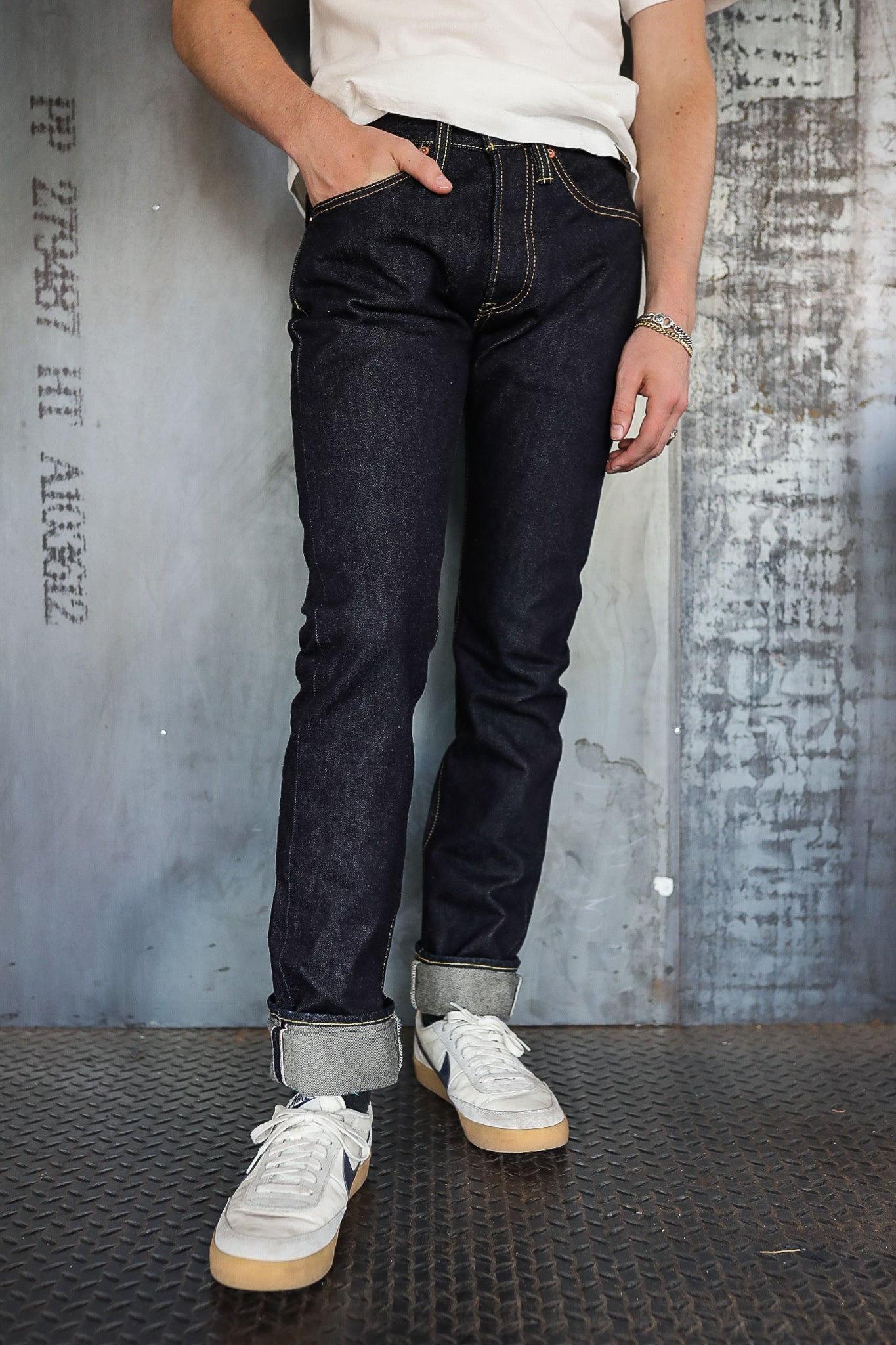 Person wearing dark raw denim jeans rolled up at cuffs with white sneakers on industrial metal floor