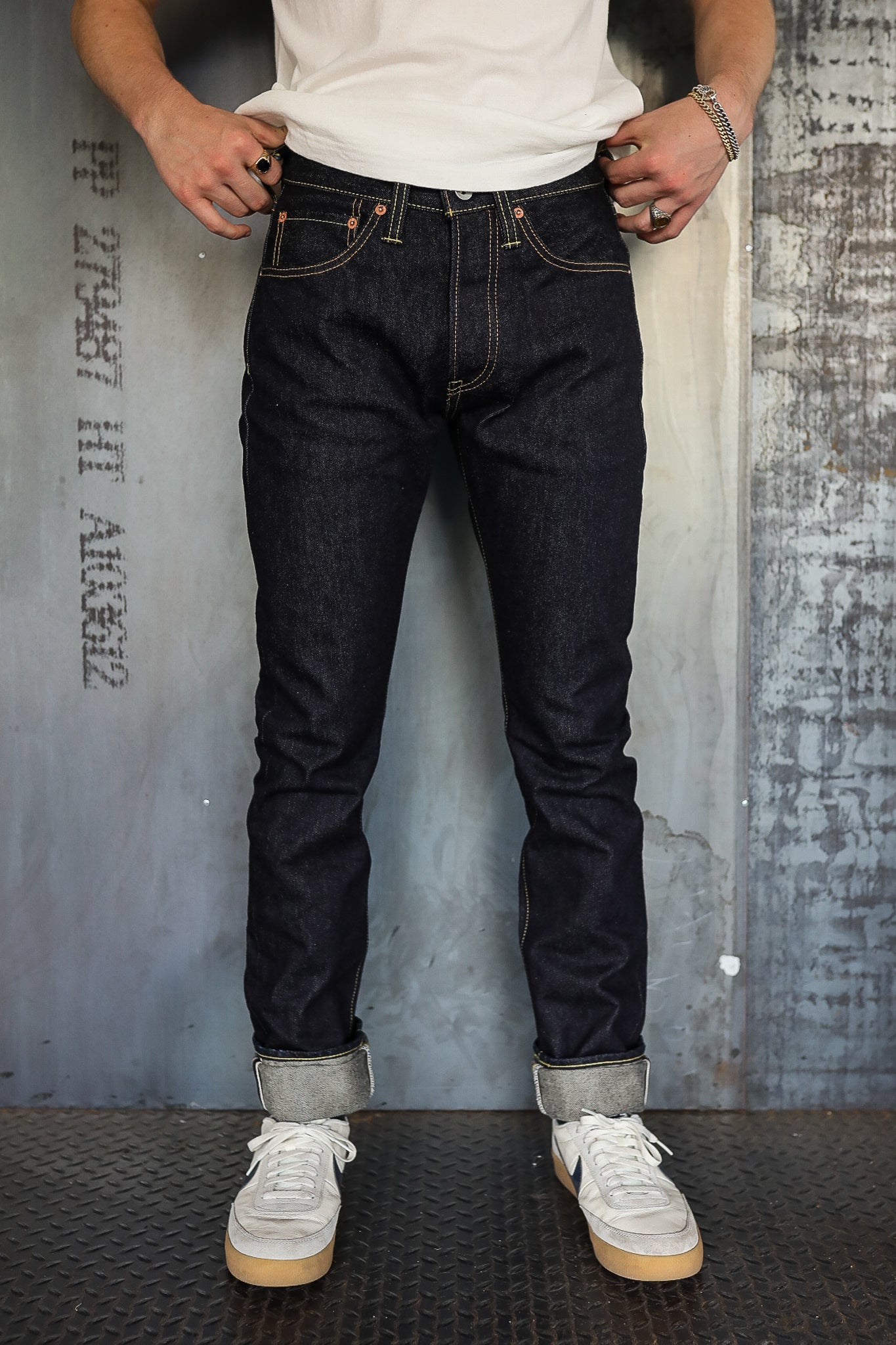 Man wearing dark raw denim jeans with rolled cuffs and white sneakers standing indoors