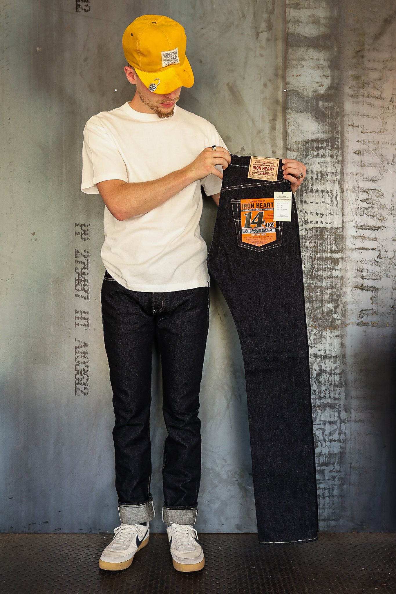 Man in white t-shirt and yellow cap holding raw selvedge denim jeans with Iron Heart 14oz label against industrial concrete wall
