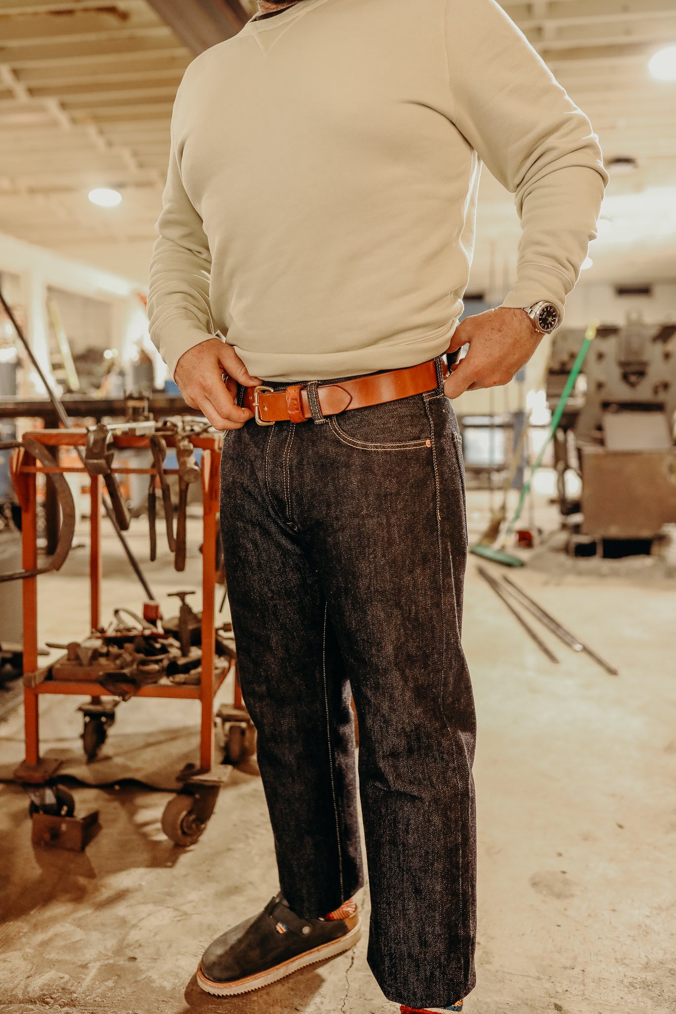 Man wearing beige sweatshirt, dark denim jeans, brown leather belt, and black slip-on shoes in workshop