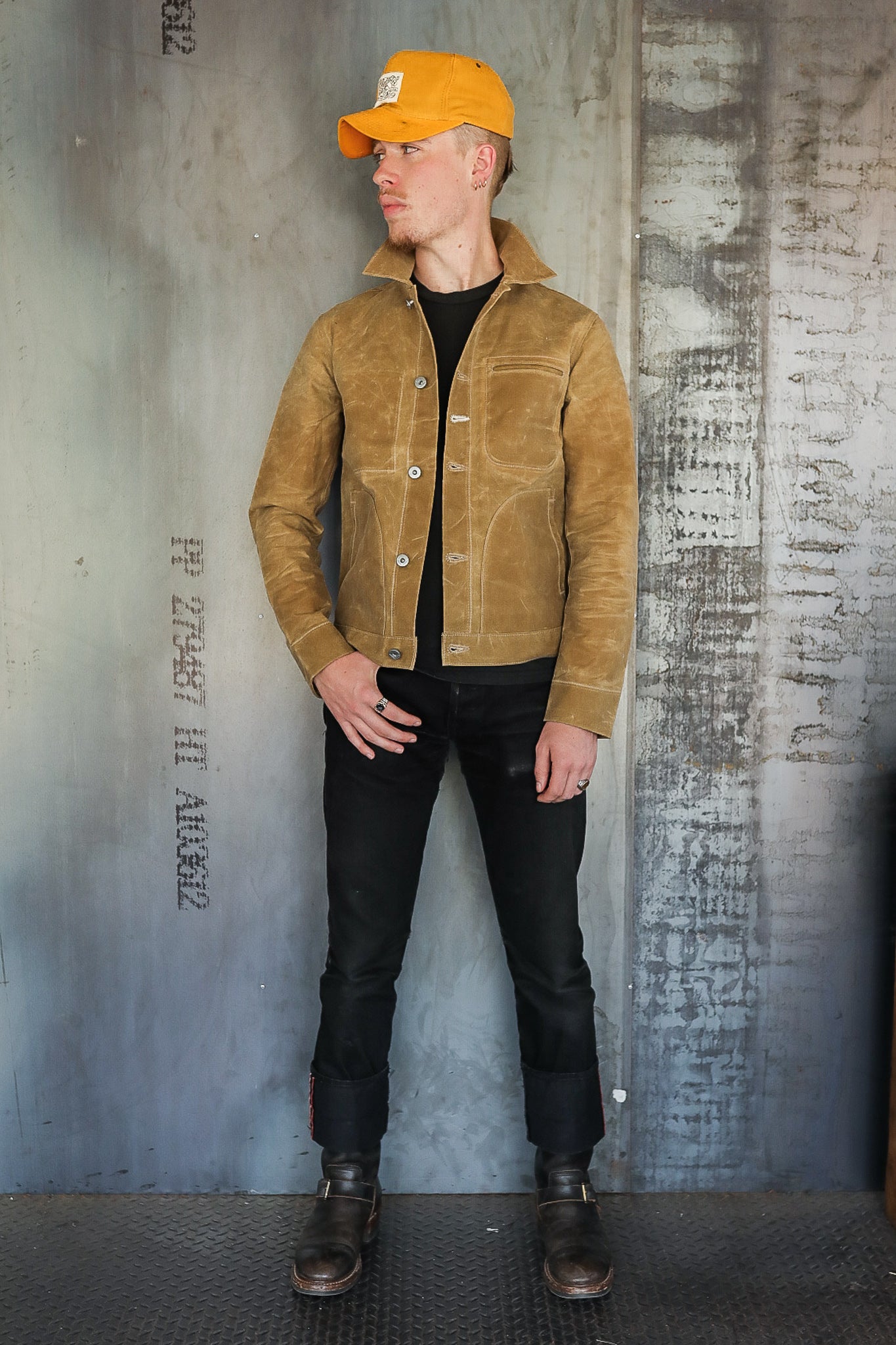 Man wearing brown leather jacket, black jeans, black boots, and yellow cap in industrial setting