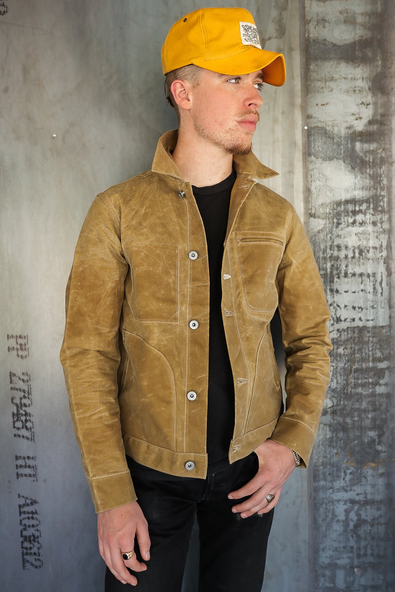 Man wearing mustard yellow canvas jacket and matching cap against industrial concrete wall at Iron Shop Provisions