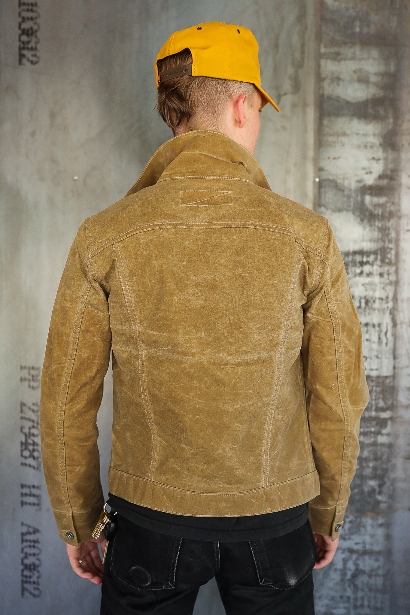Man wearing tan waxed canvas work jacket and yellow cap, standing against textured industrial wall