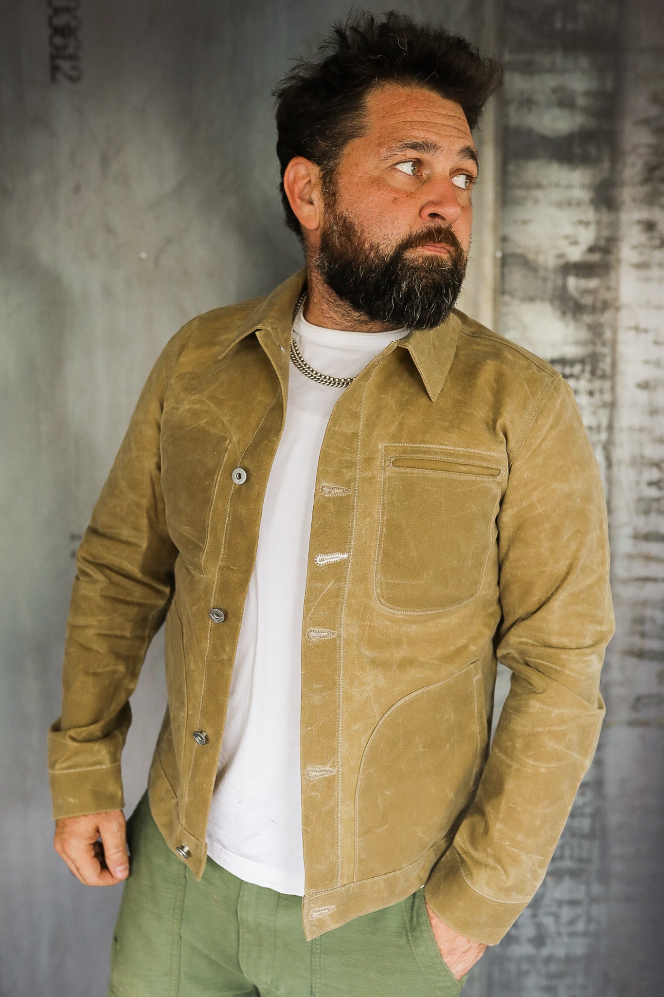 Man wearing tan waxed canvas chore jacket with green pants and white shirt against industrial metal background