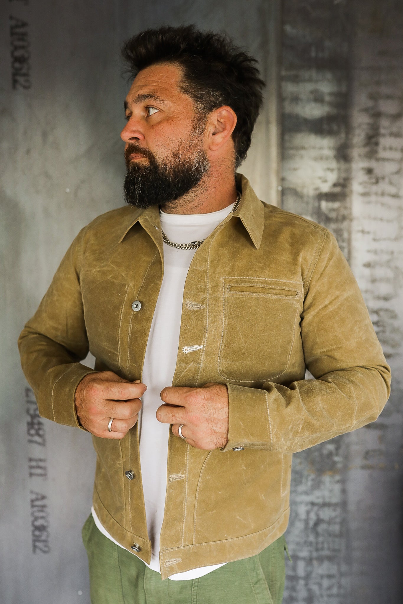 Man with beard wearing a tan waxed canvas jacket over white shirt and green pants against industrial background