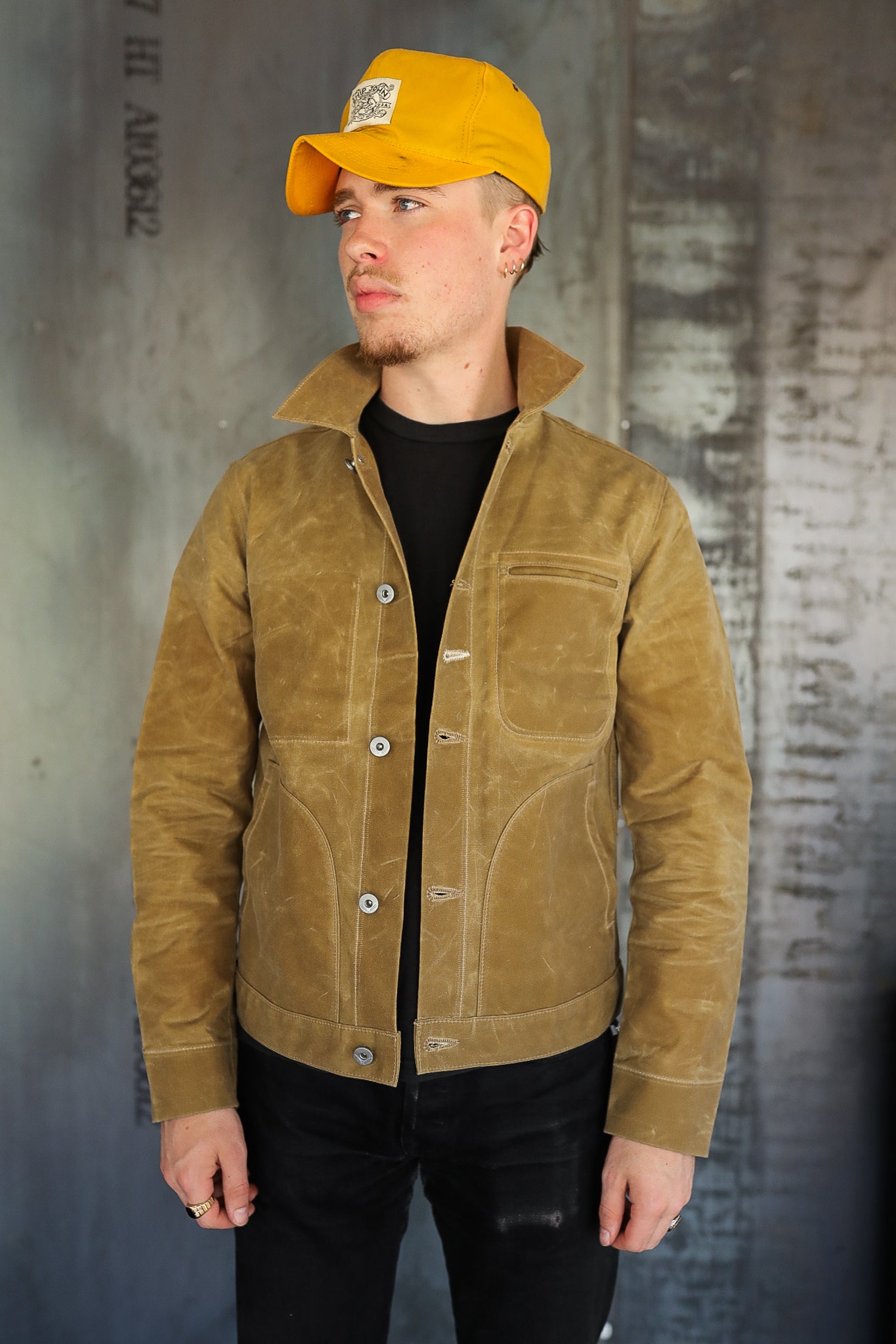 man wearing tan waxed canvas jacket and yellow cap standing against industrial background