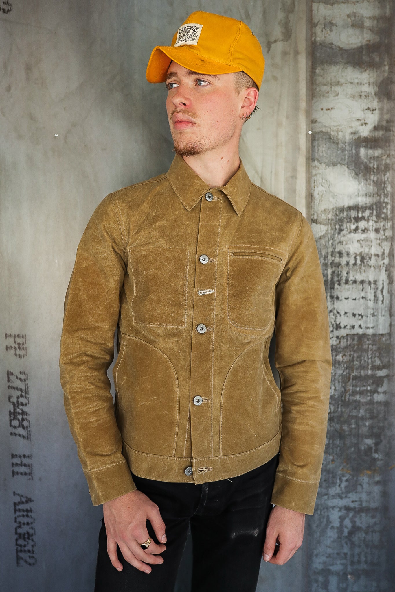 Man wearing tan waxed canvas jacket and mustard yellow cap against industrial wall