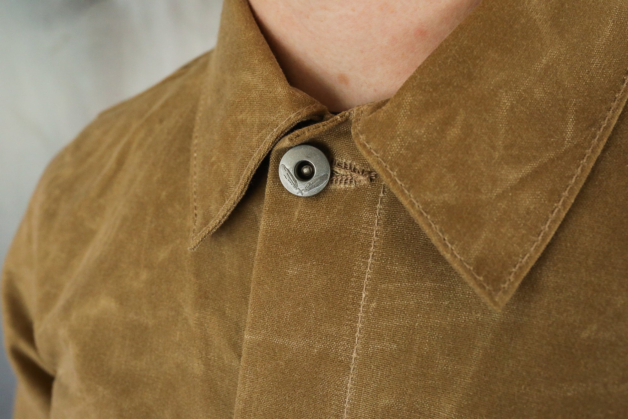 Close-up of brown canvas jacket collar with metal button featuring crossed feather design