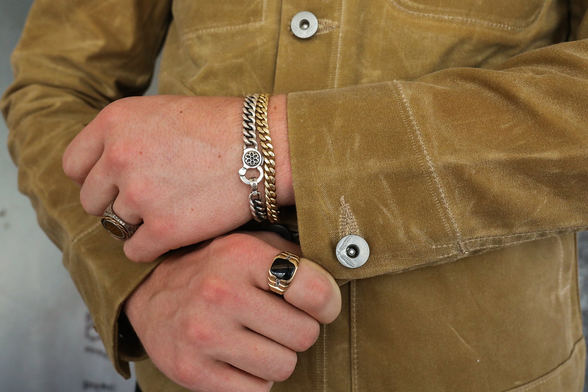 Close-up of hands wearing gold and silver chain bracelets and rings with black stone, styled with brown work jacket