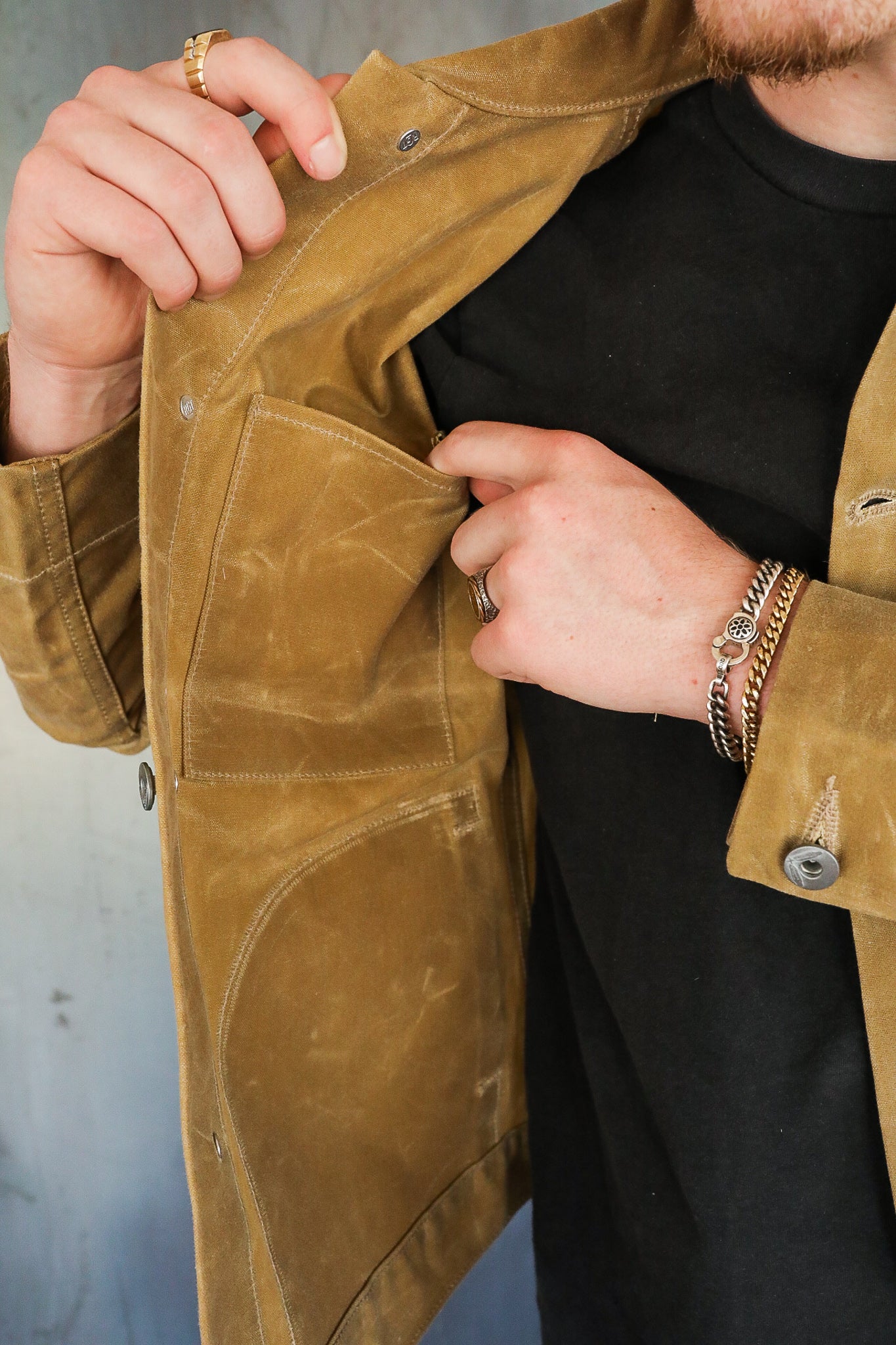 Close-up of a person wearing a tan rugged jacket showing the inside pocket, dressed in black shirt with metal bracelets and rings