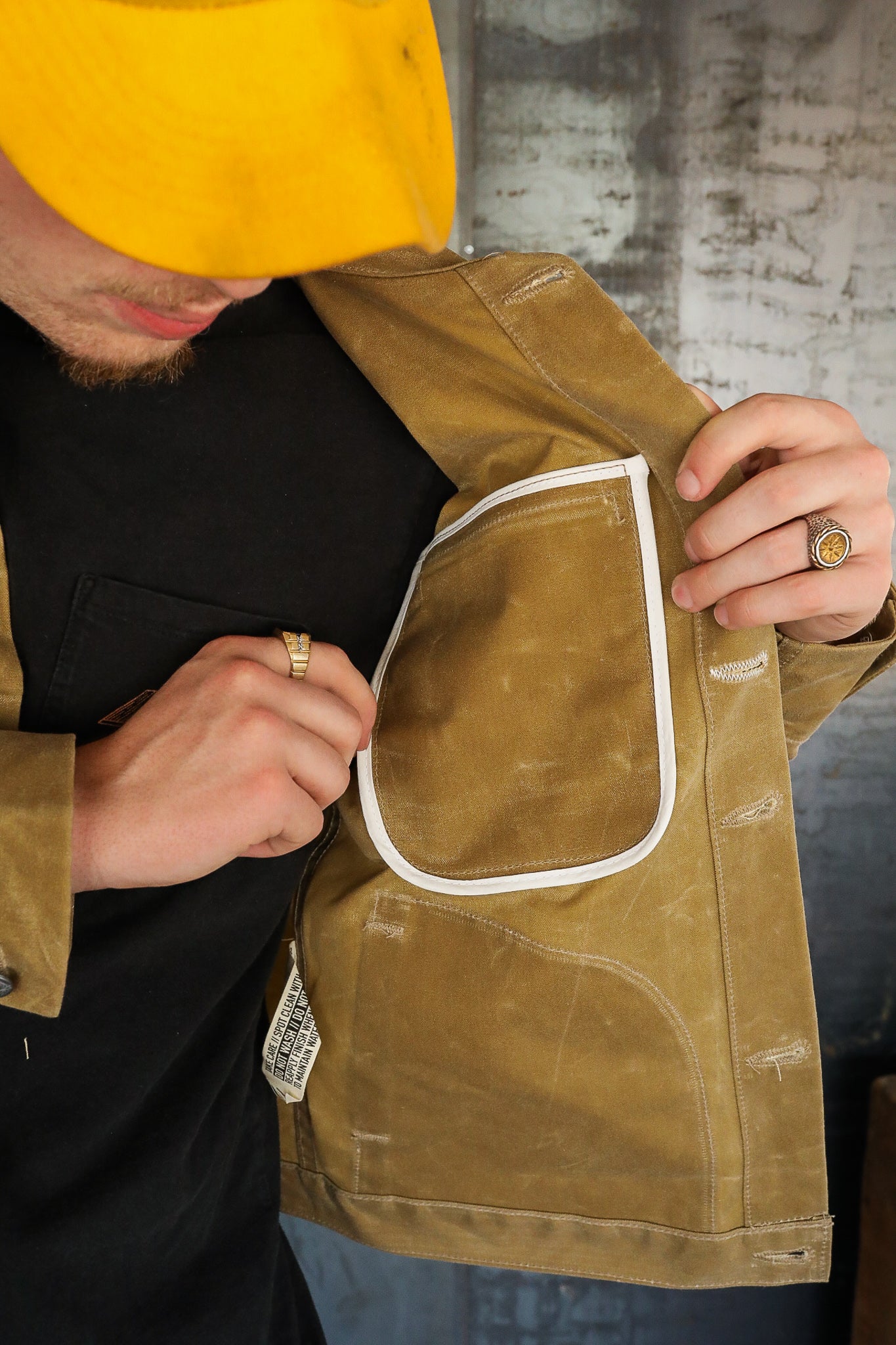 Man wearing yellow cap and brown jacket showing interior pocket detail with white trim at Iron Shop Provisions