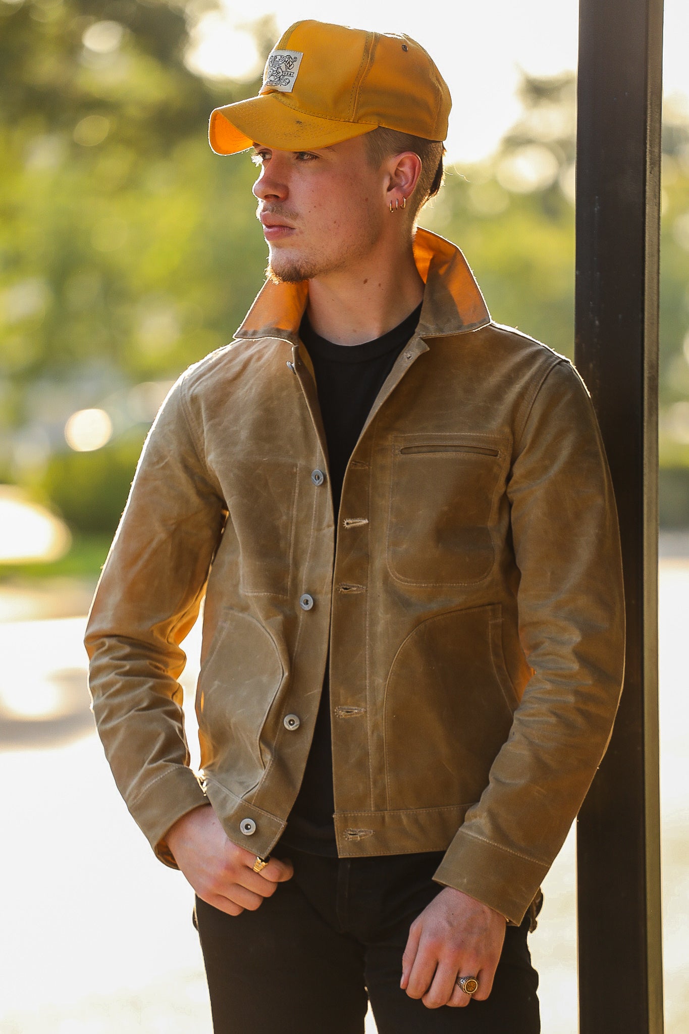 Young man wearing tan leather jacket and yellow cap with Iron Shop Provisions patch outdoors