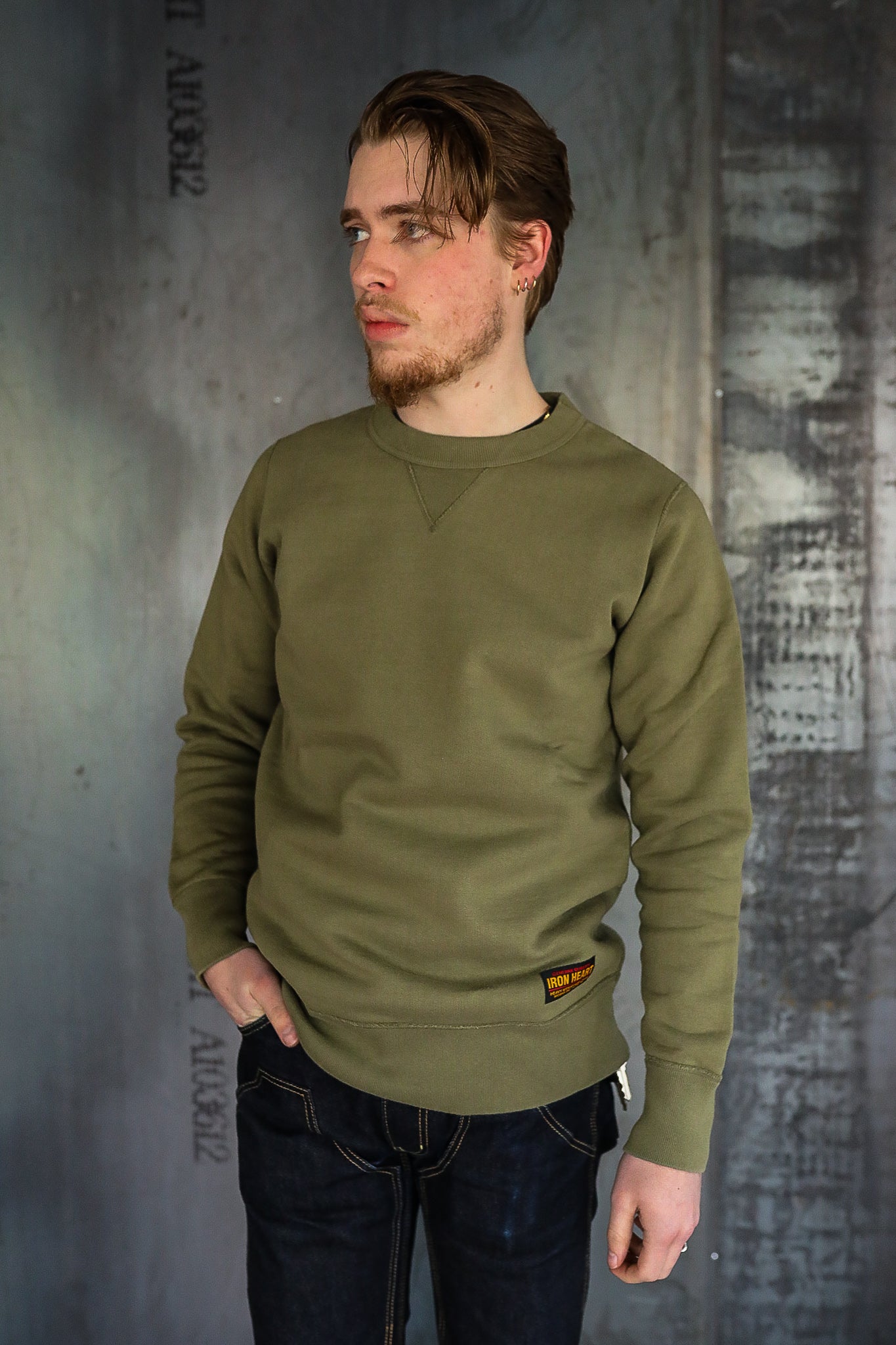 Man wearing olive green Iron Heart sweatshirt and dark denim jeans against industrial gray wall