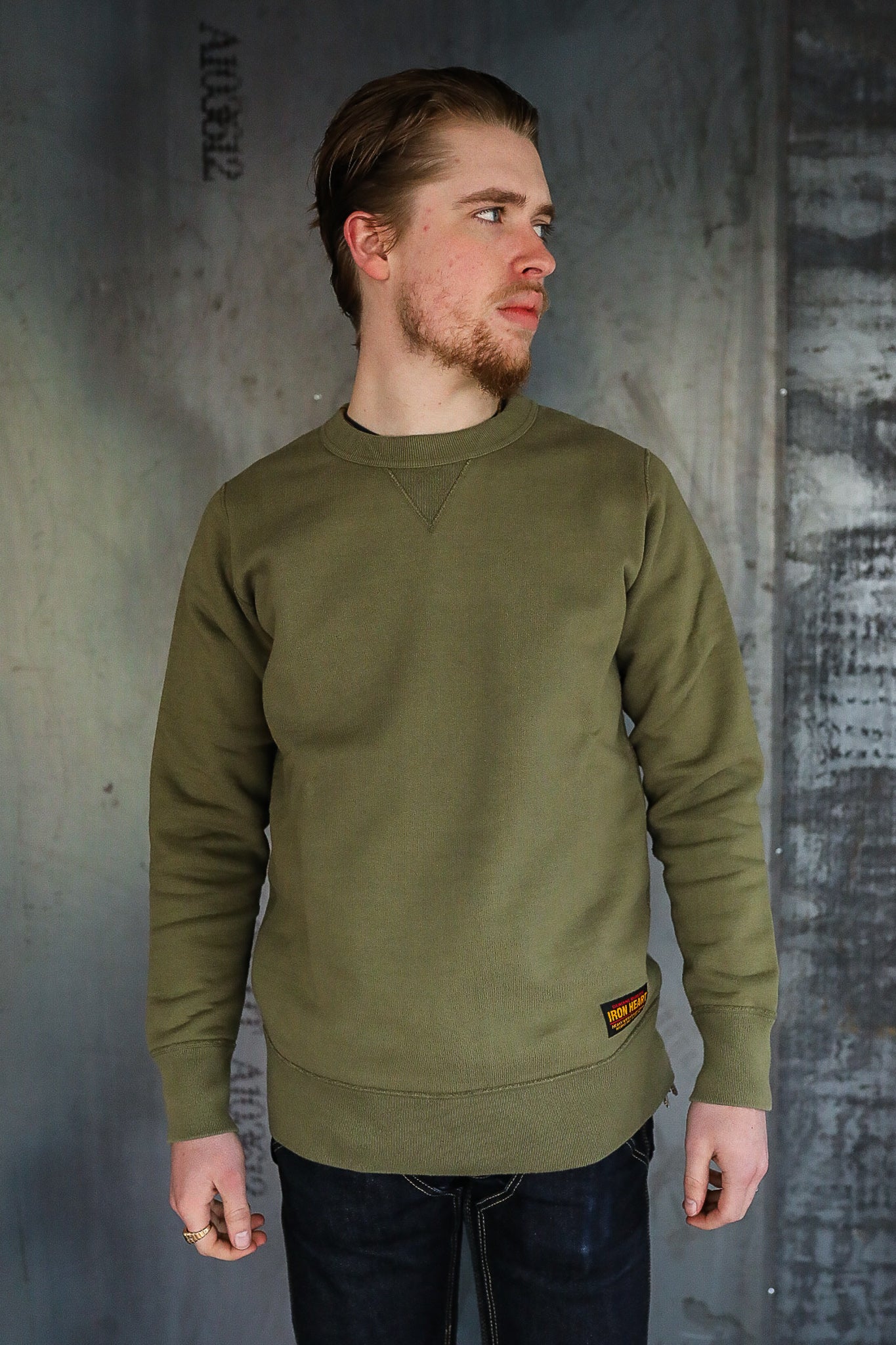 Man wearing olive green Iron Heart sweatshirt and dark jeans against industrial metal background