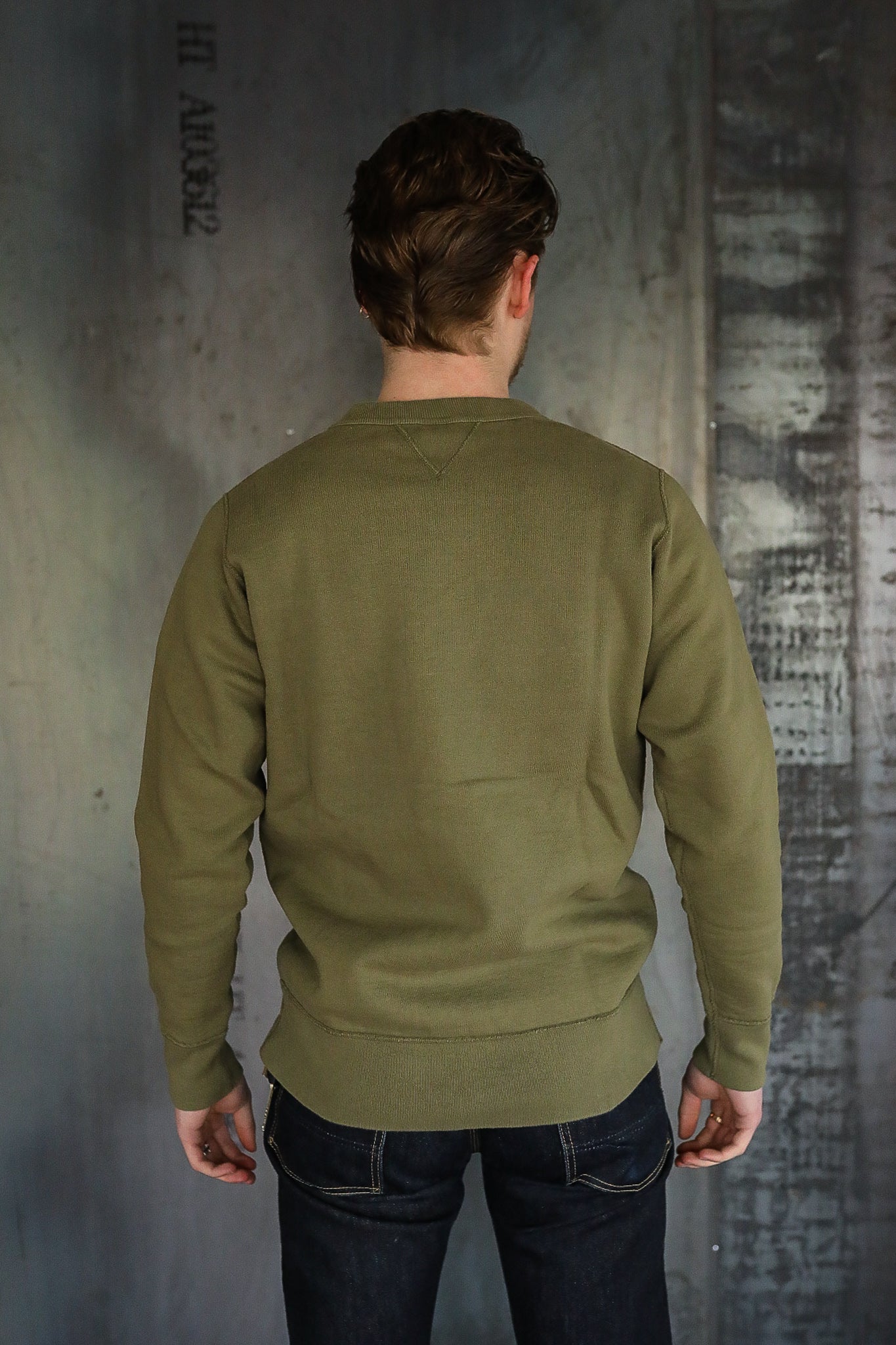 Man wearing olive green sweatshirt and dark denim jeans standing with back to camera against industrial wall