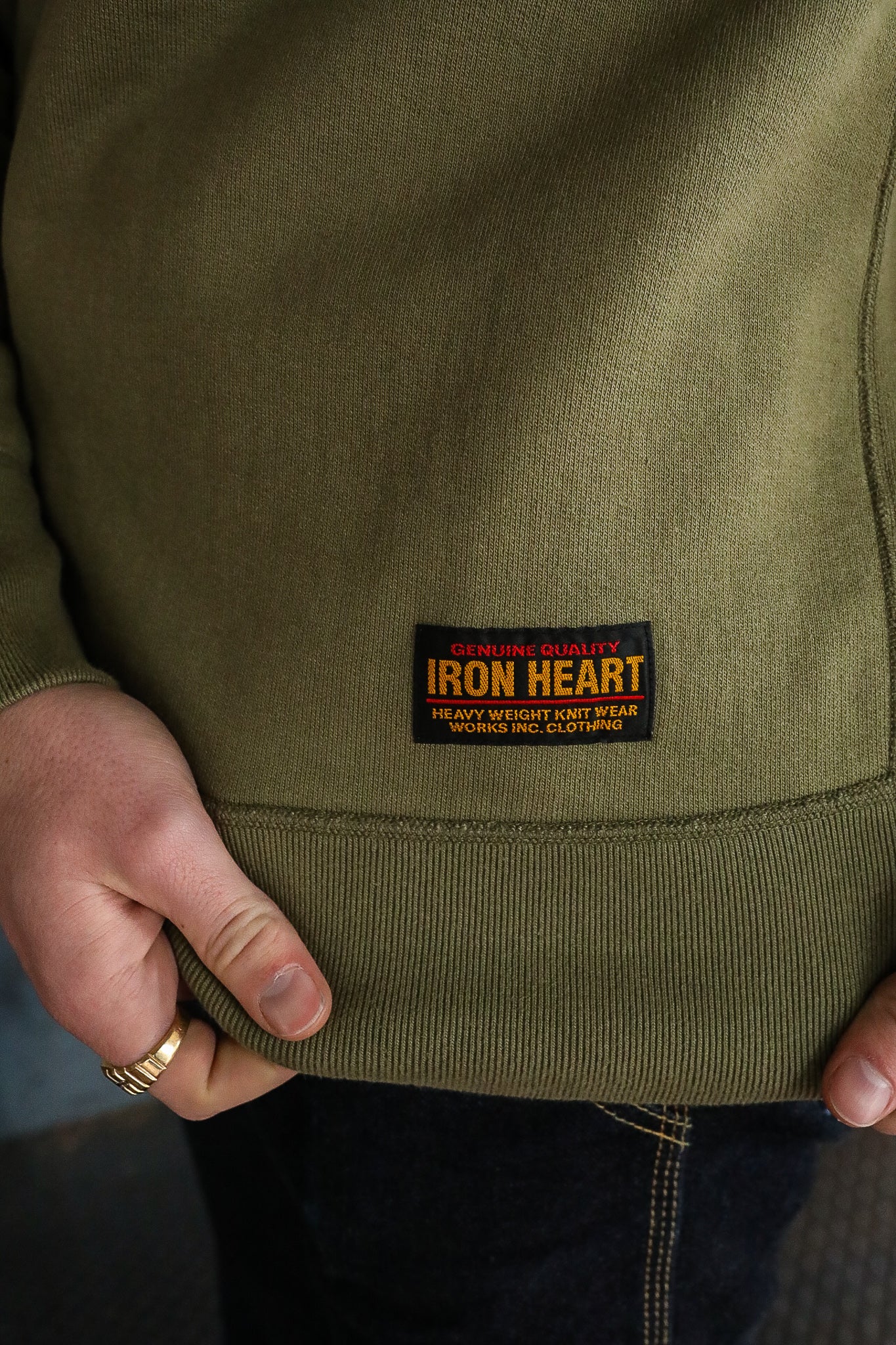Close-up of olive green Iron Heart sweatshirt with label and hand wearing gold ring, heavy knitwear