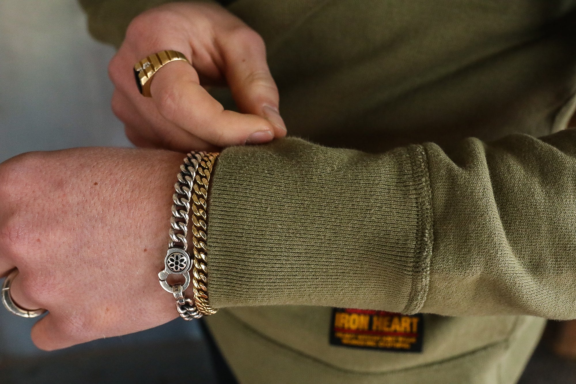 Close-up of a person's wrist wearing silver and gold chain bracelets with a green Iron Heart sweatshirt sleeve