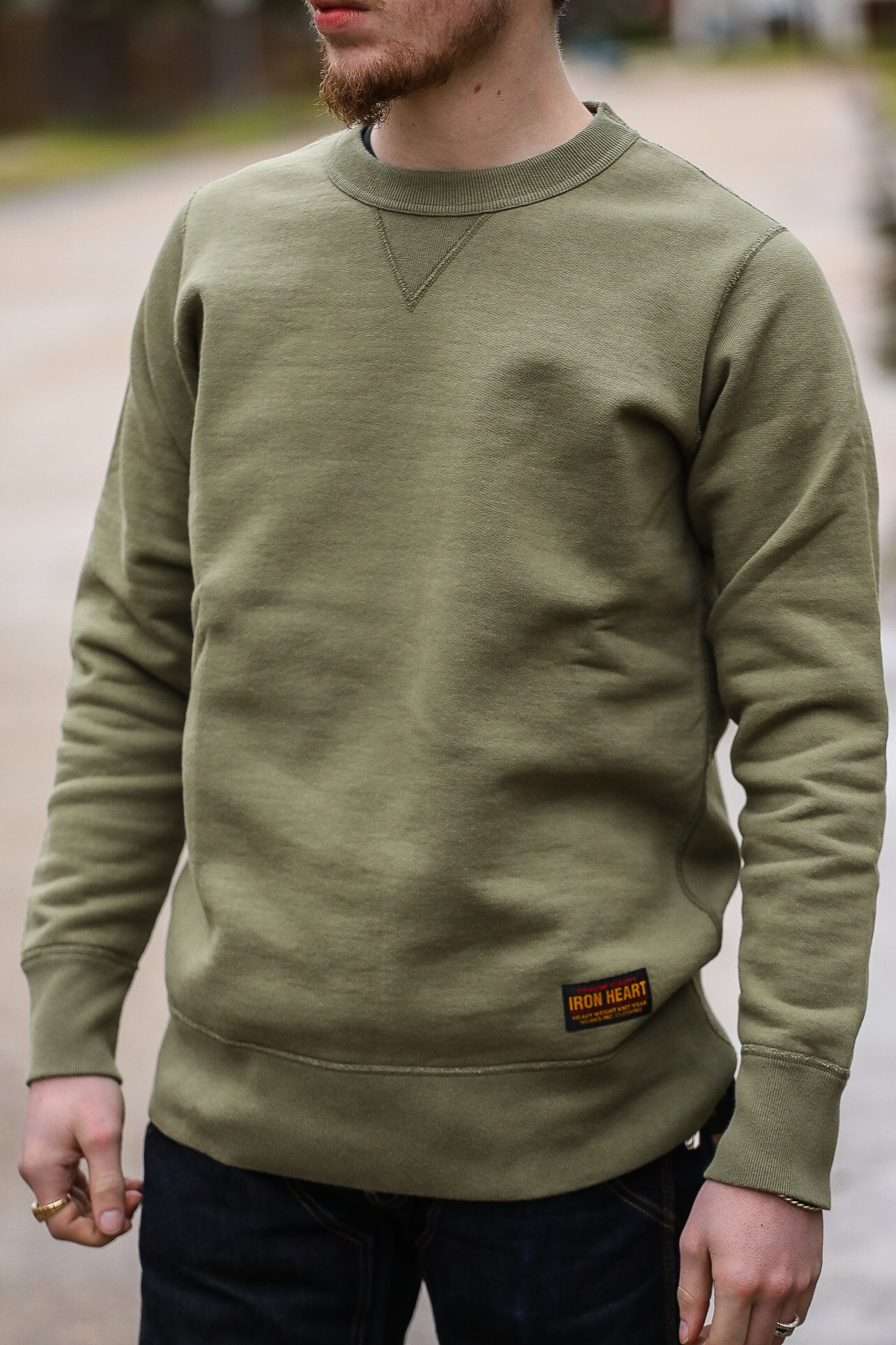 Man wearing olive green Iron Heart crewneck sweatshirt with ribbed cuffs and hem, outdoor setting