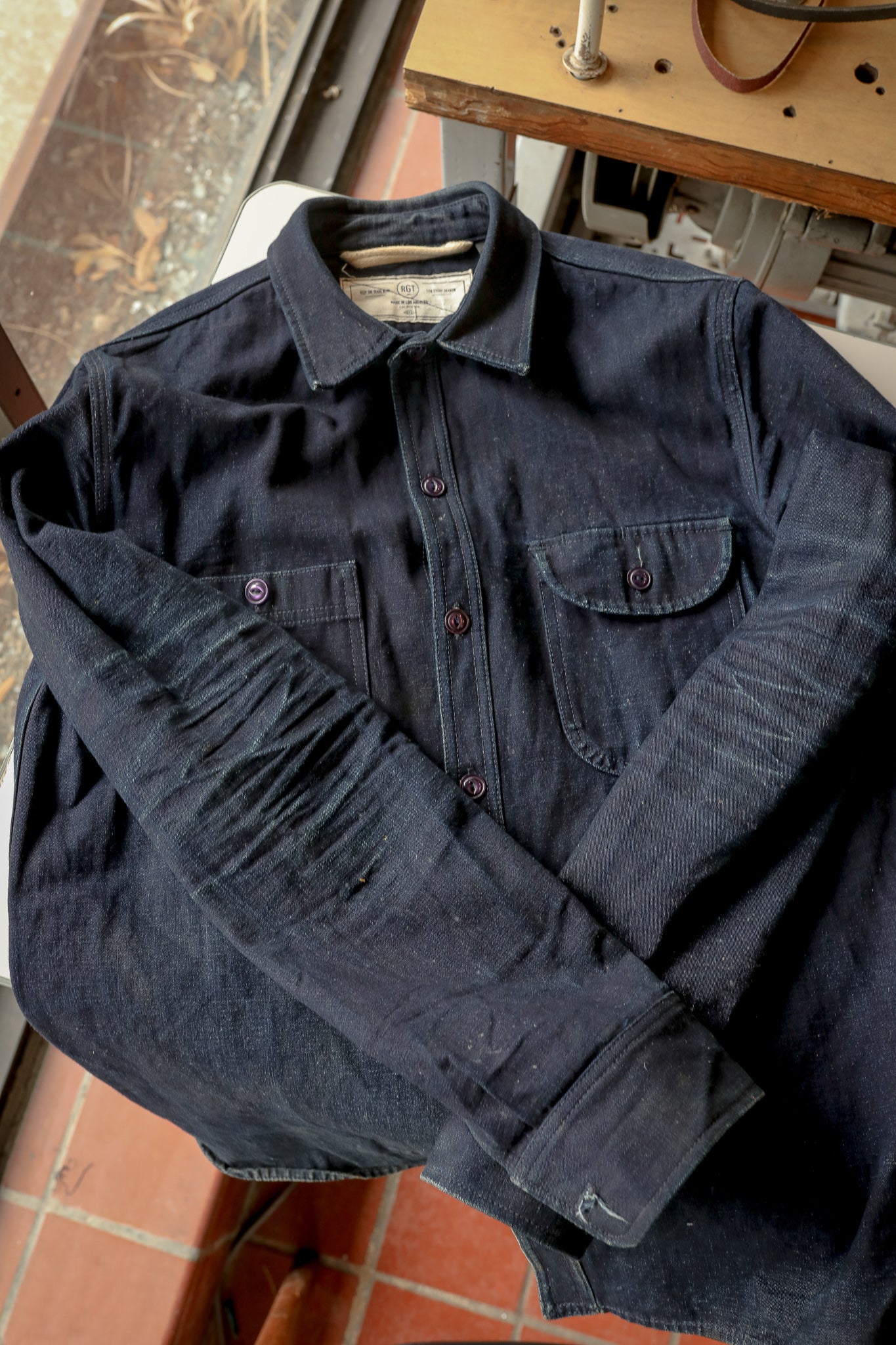 Dark indigo denim chore jacket with front pockets and buttoned cuffs laid on chair by window