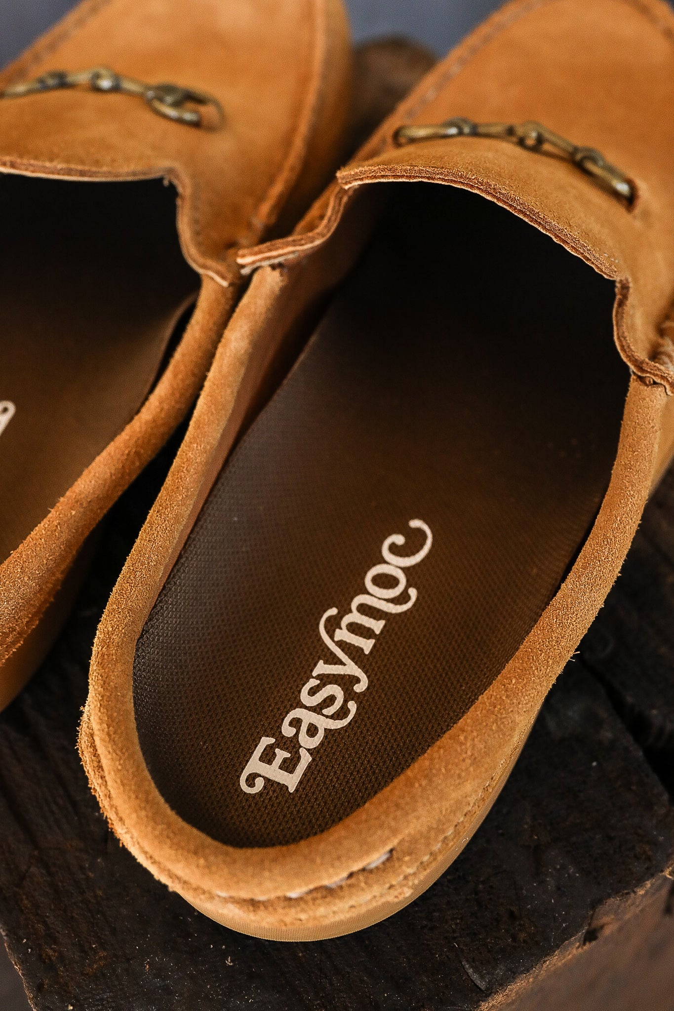 Close-up of tan suede Easy Moc slip-on casual shoes with metal bit detail on dark wood surface