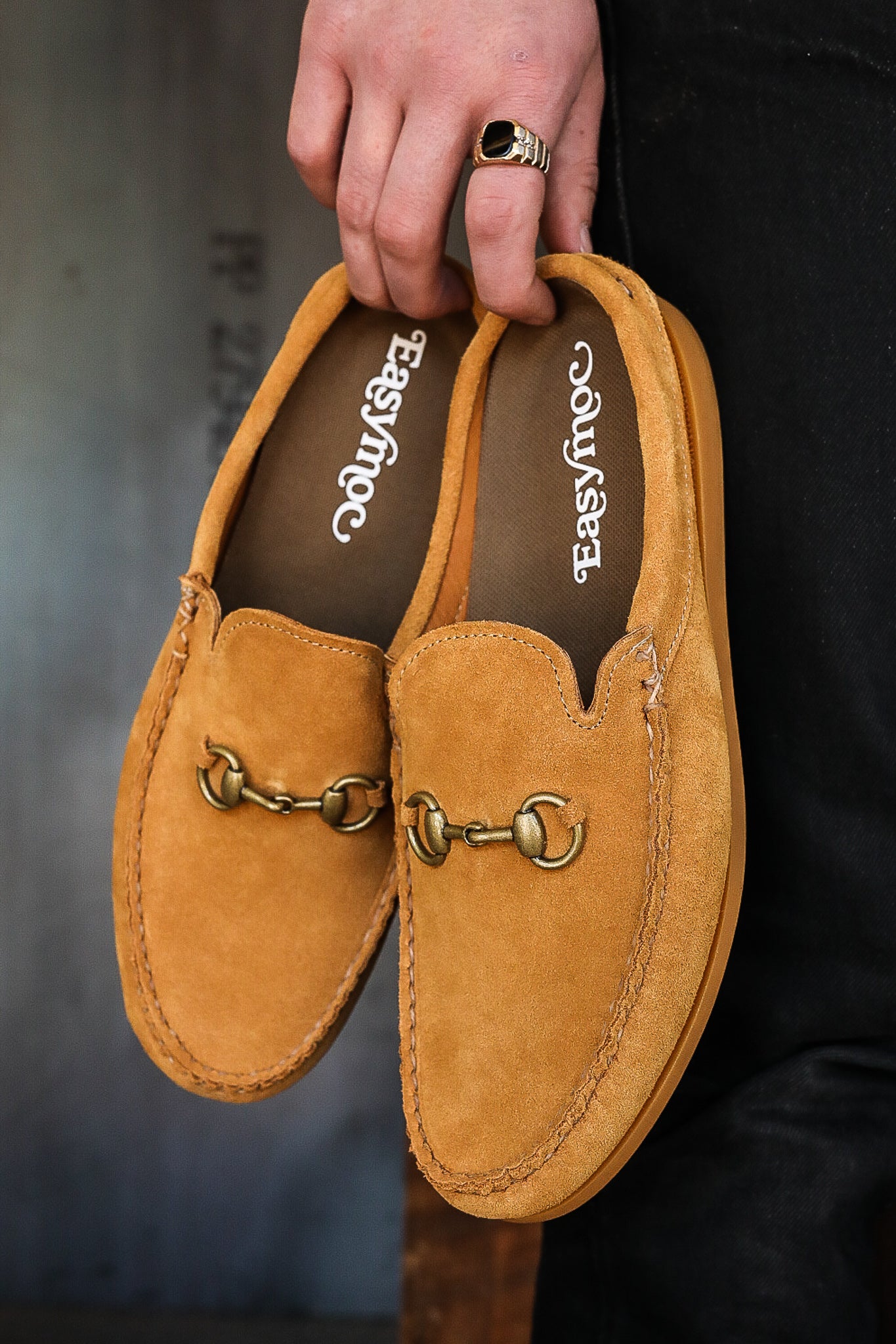 Hand holding pair of tan suede Easy Moc loafers with metal bit detail and stitched trim
