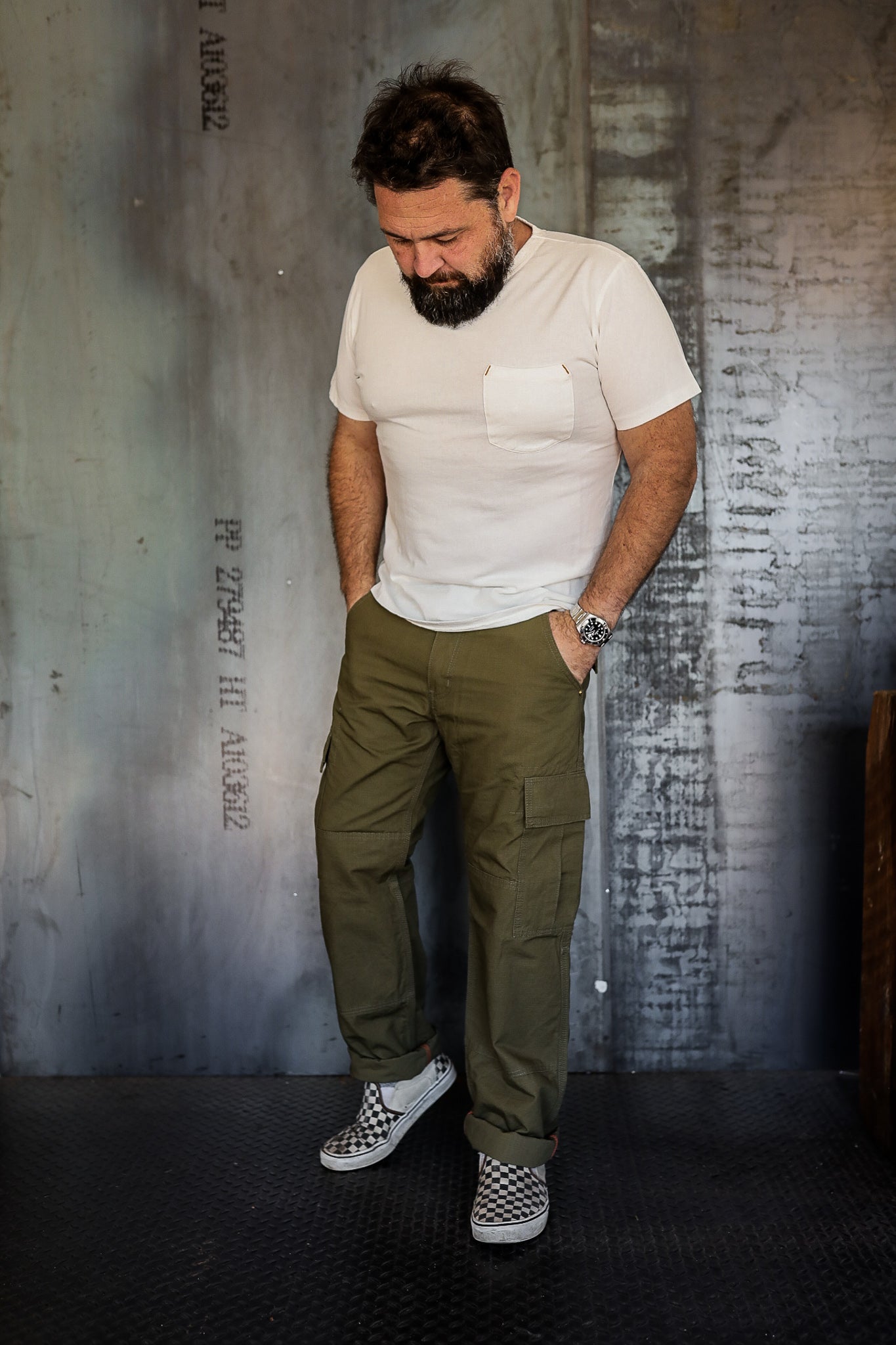 Man wearing white pocket t-shirt, olive cargo pants, and checkered slip-on shoes in industrial setting