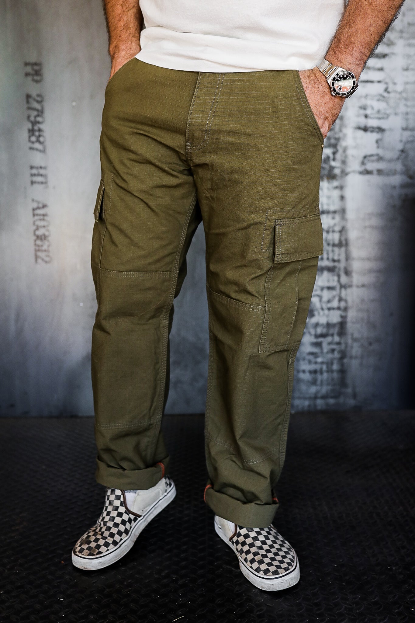 man wearing olive green cargo pants and black-and-white checkered slip-on sneakers in industrial setting