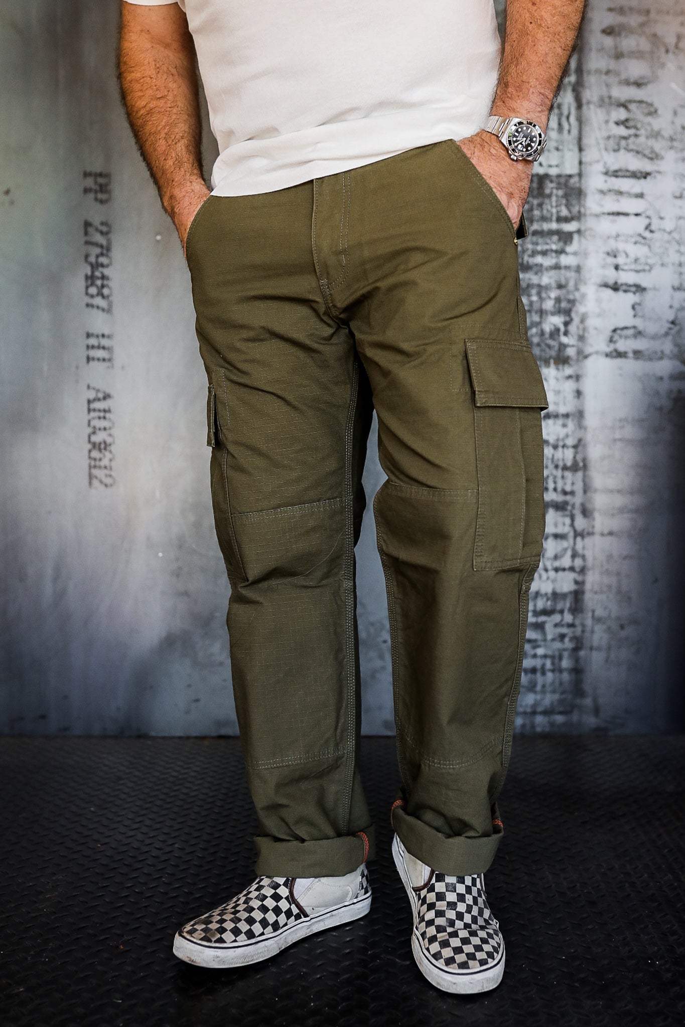 Man wearing olive green cargo pants with checkered slip-on sneakers against industrial metal background