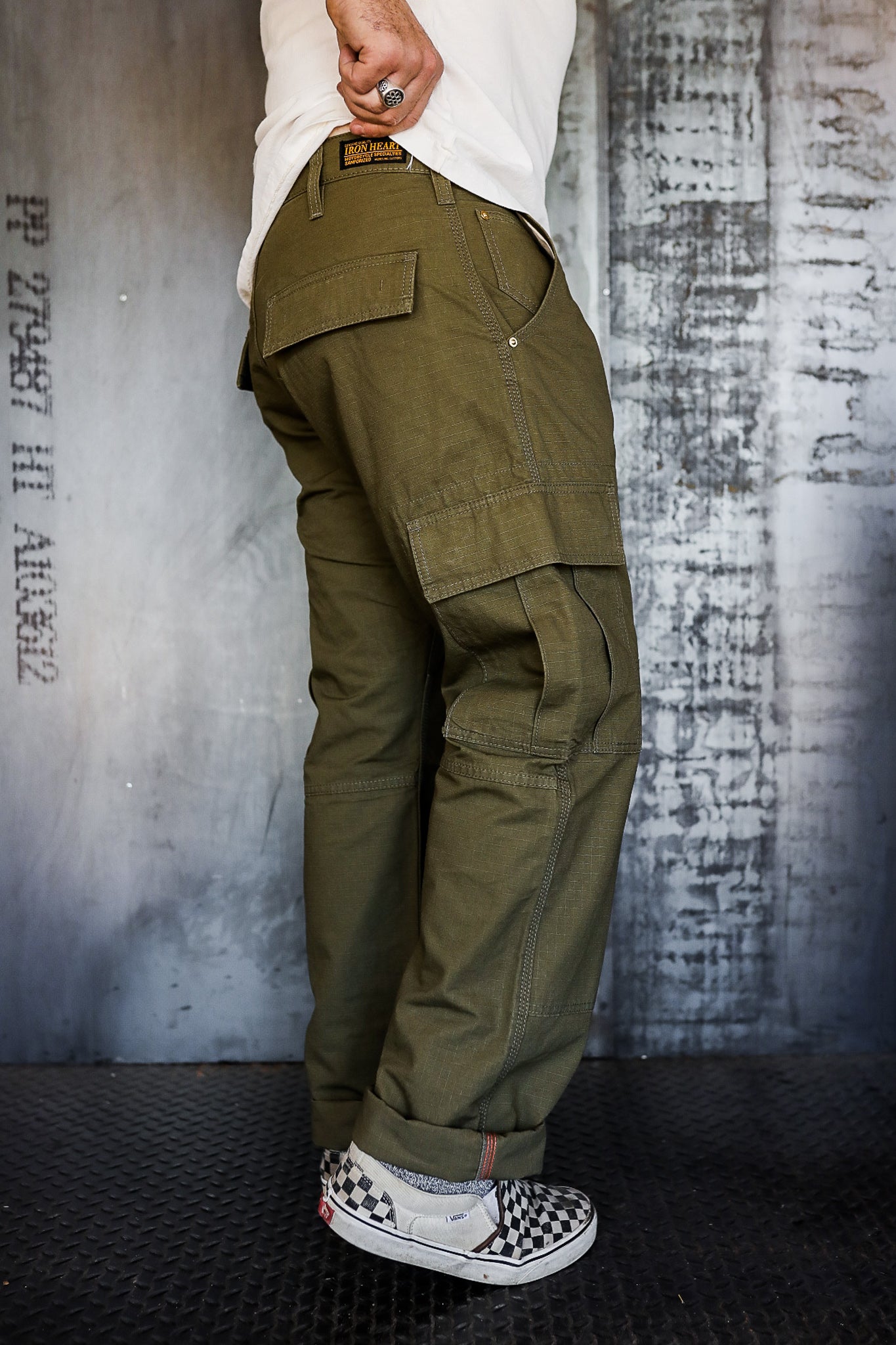 Close-up of man wearing olive green cargo pants with multiple pockets and sneakers against industrial wall