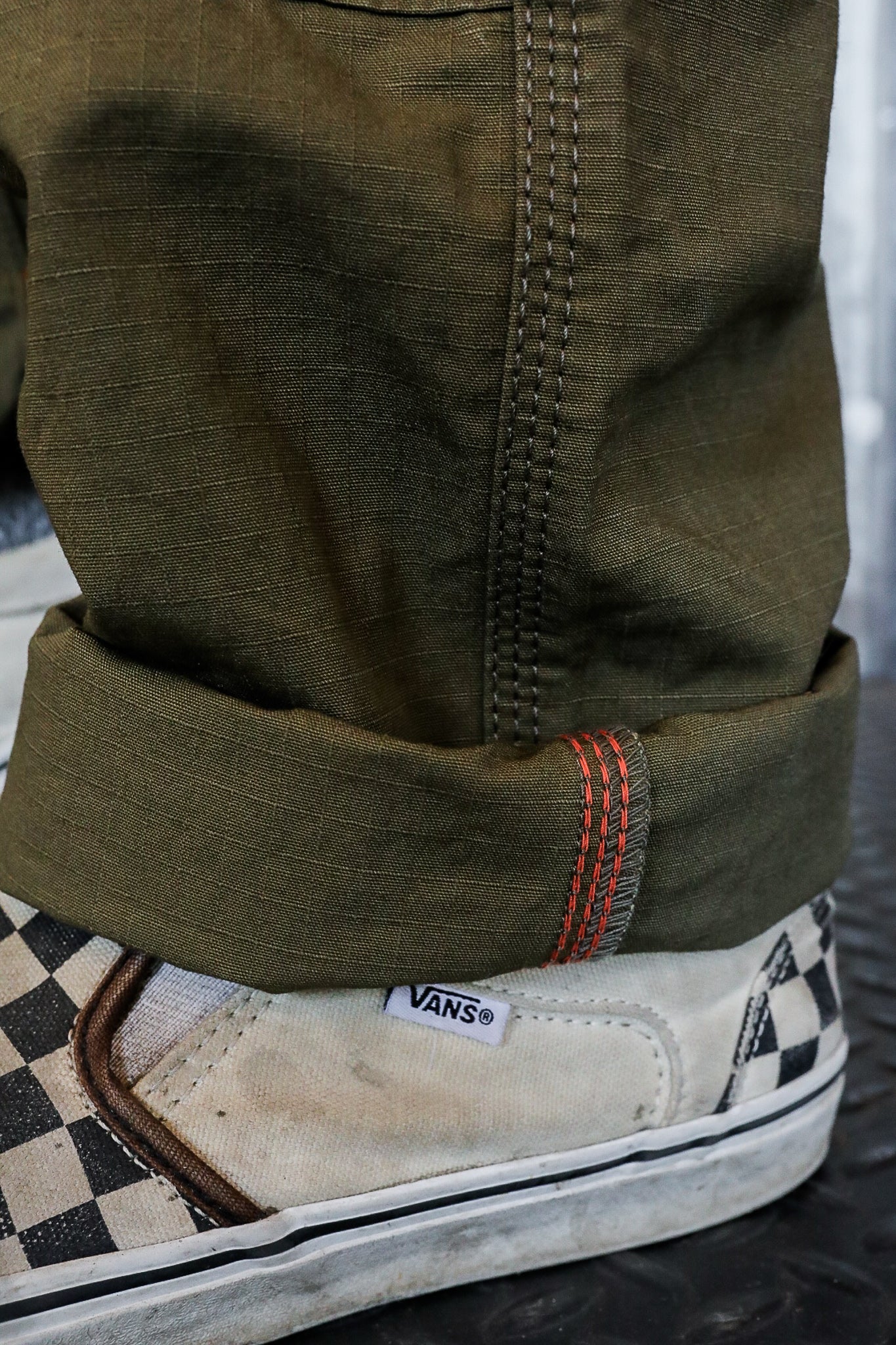 Close-up of olive green work pants cuff with orange stitching over worn white Vans checkered slip-on sneakers