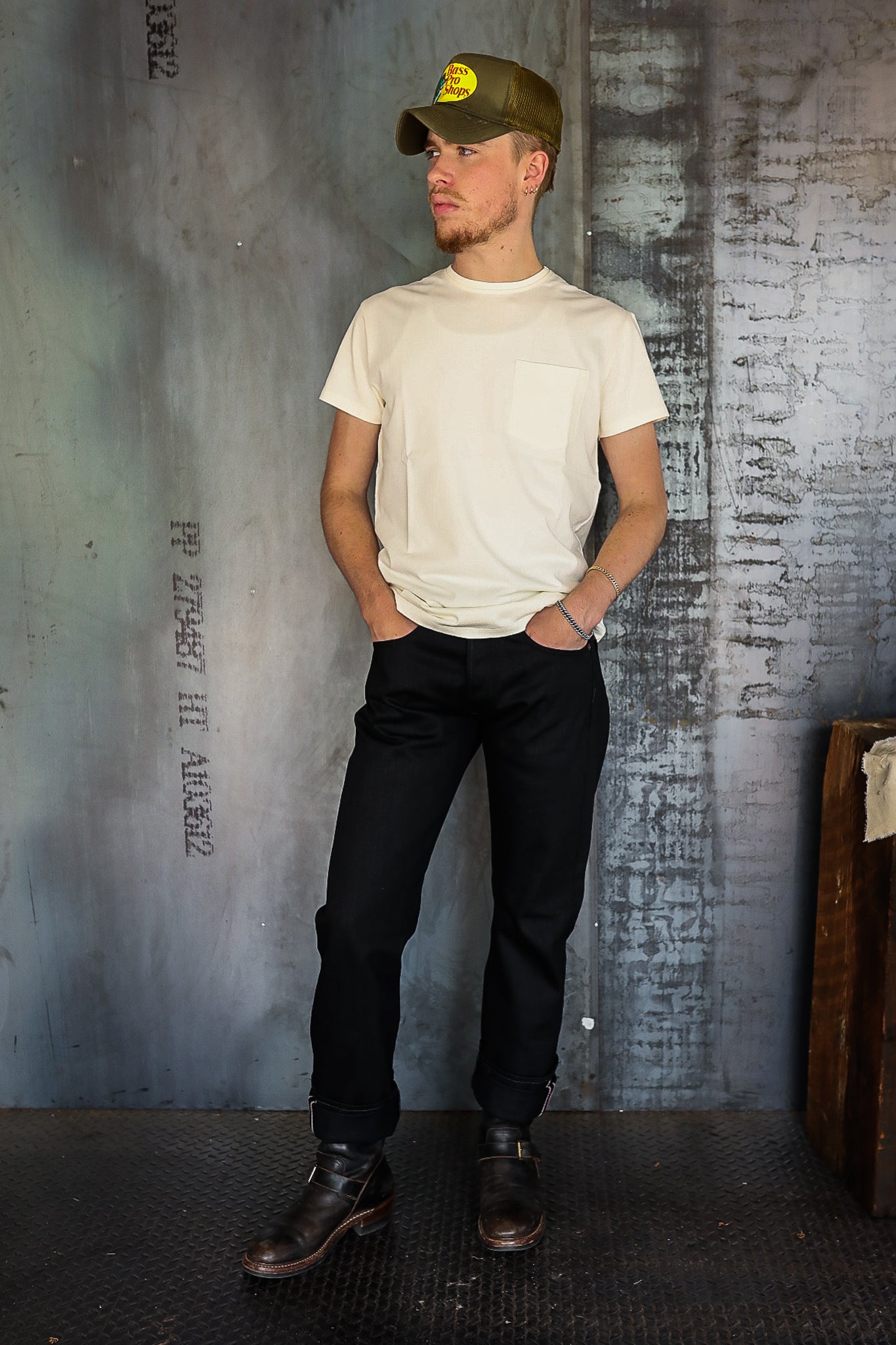 Man wearing cream pocket t-shirt, black jeans, dark leather boots, and olive green Bass Pro Shops cap in industrial setting