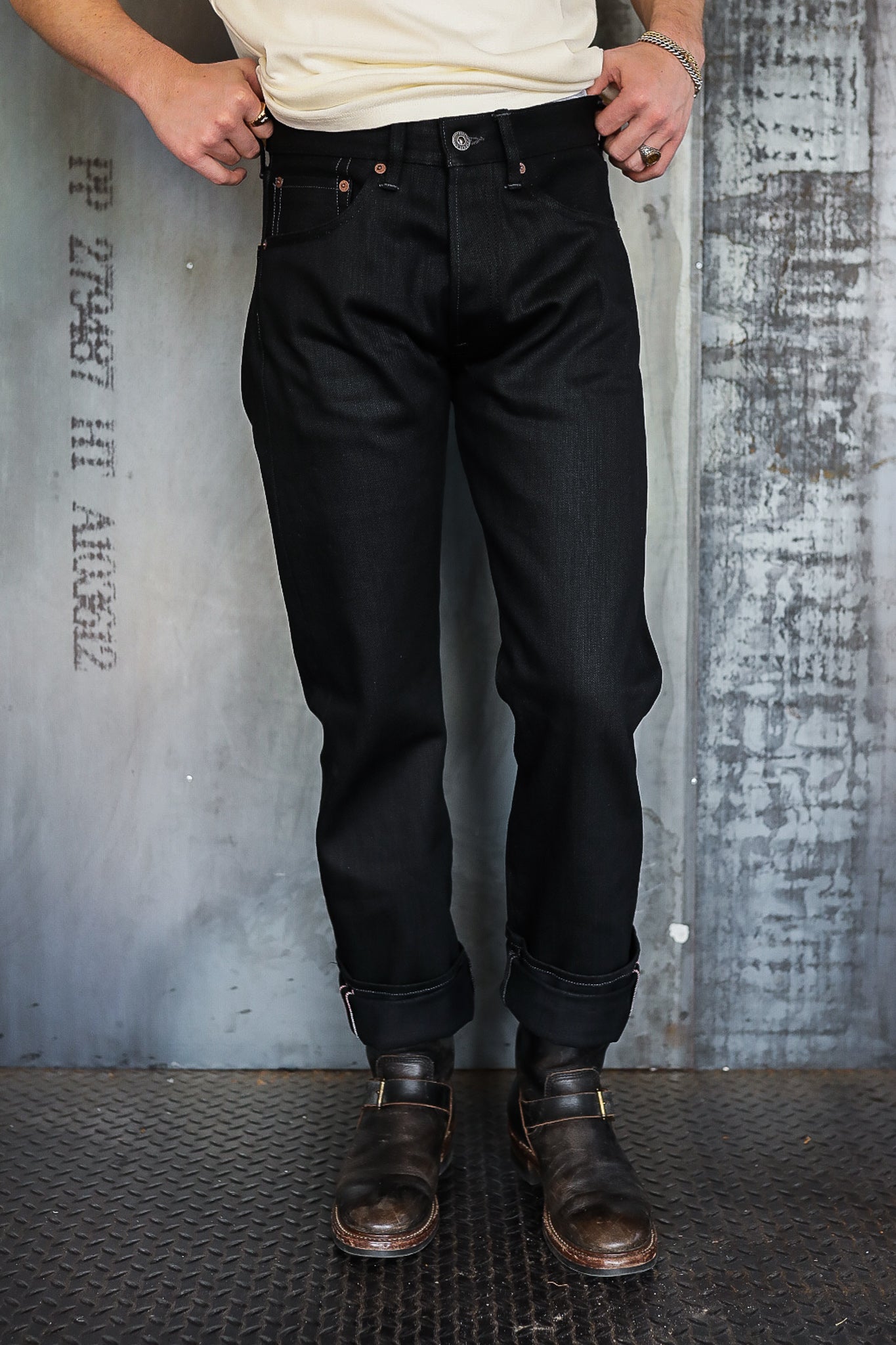 Person wearing black selvedge denim jeans with cuffed hems and brown leather boots against industrial background