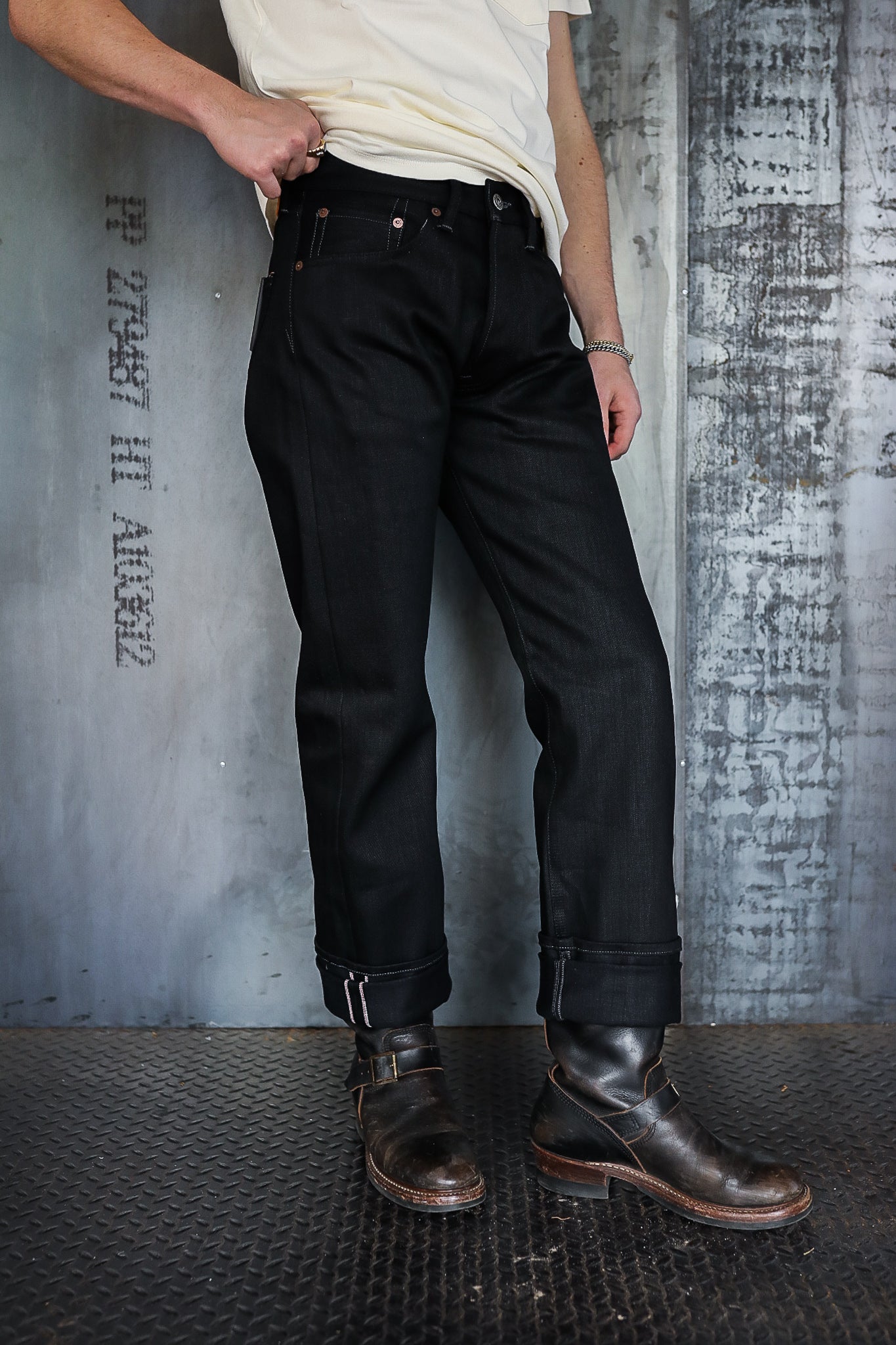 Man wearing dark raw denim jeans with cuffs and black leather boots against industrial metal background
