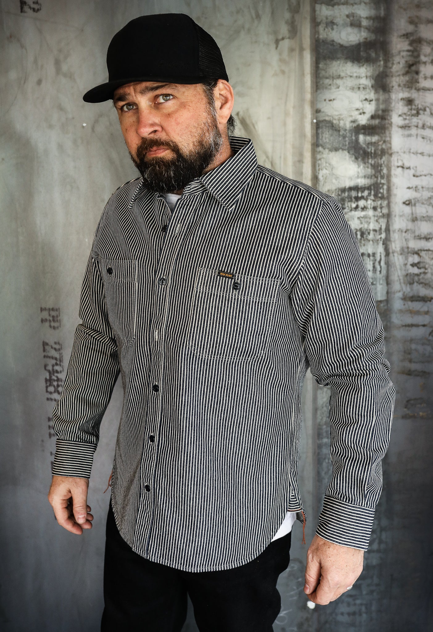 Bearded man wearing black and white striped work shirt and black cap against industrial metal background