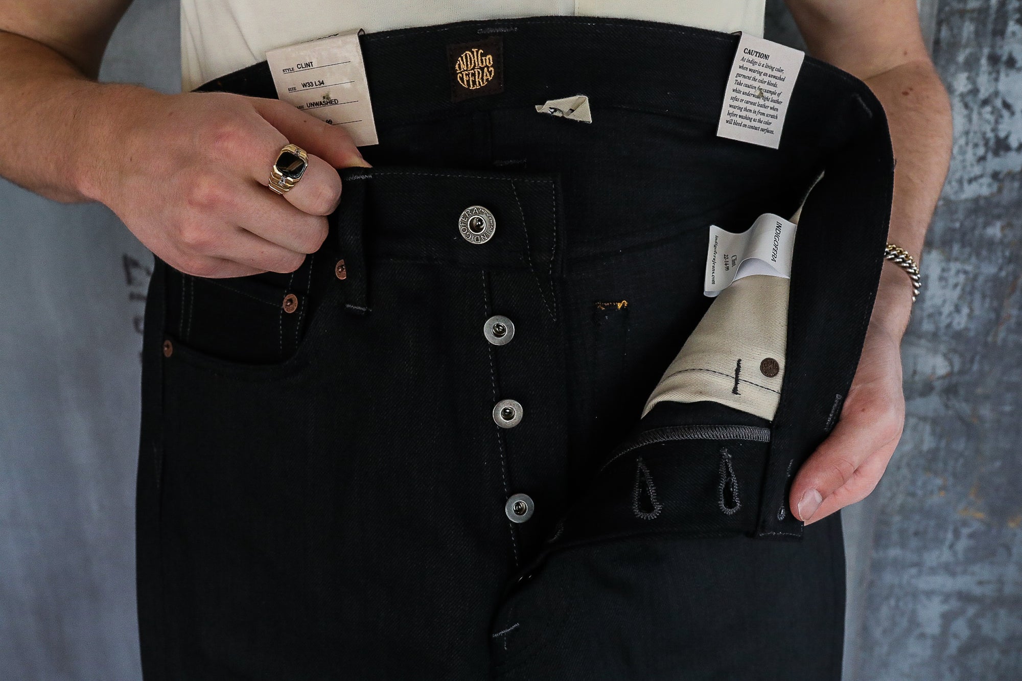 Person holding open black Indigofera Clint raw denim jeans with visible button fly, brand tags, and copper rivets