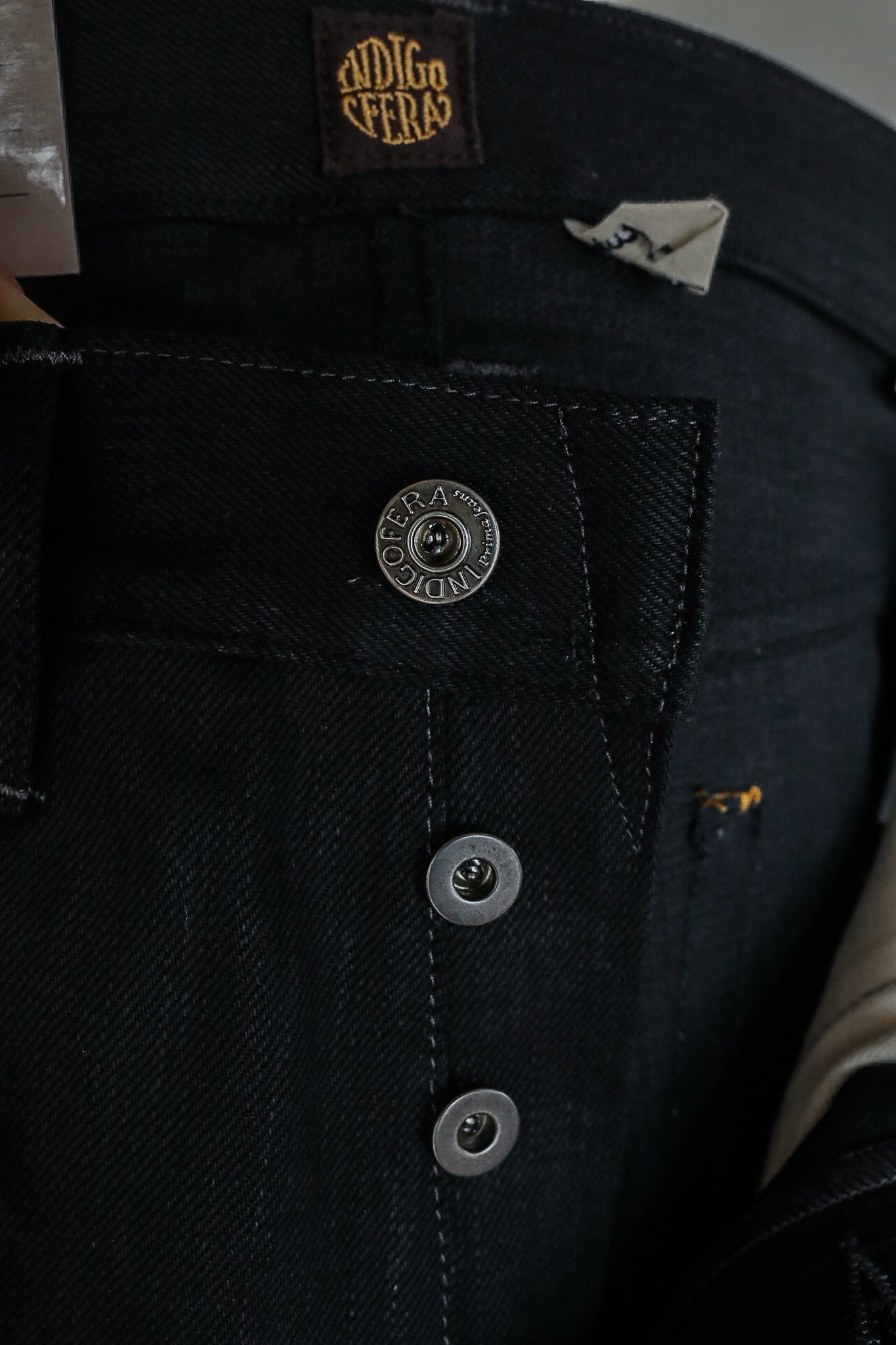 Close-up of black denim jeans with metal buttons and Indigofera brand label