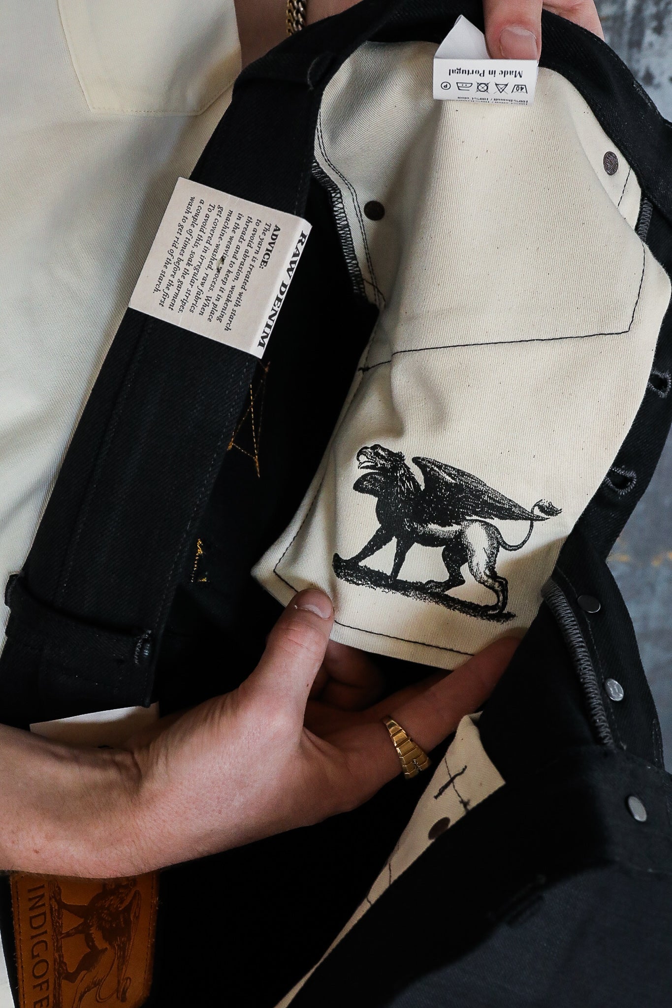 Hands holding black denim jacket showing the inside lining with a black griffin graphic and care labels