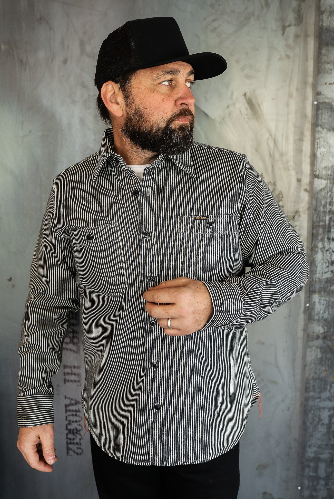 Bearded man wearing black and white striped work shirt and black cap against industrial wall