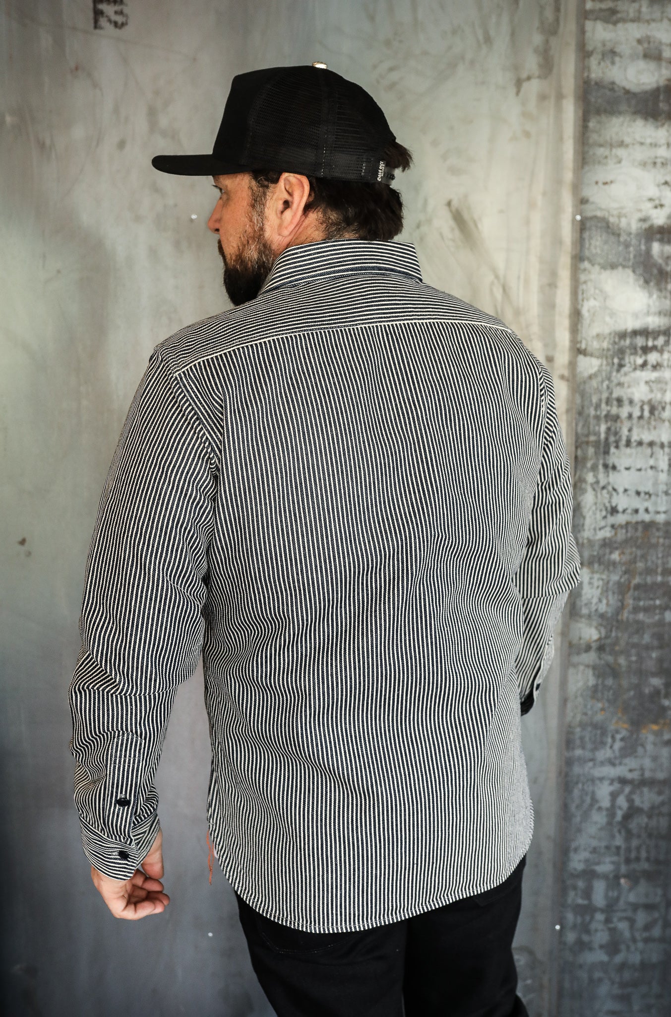 Man wearing black and white striped work shirt and black mesh cap standing in industrial setting