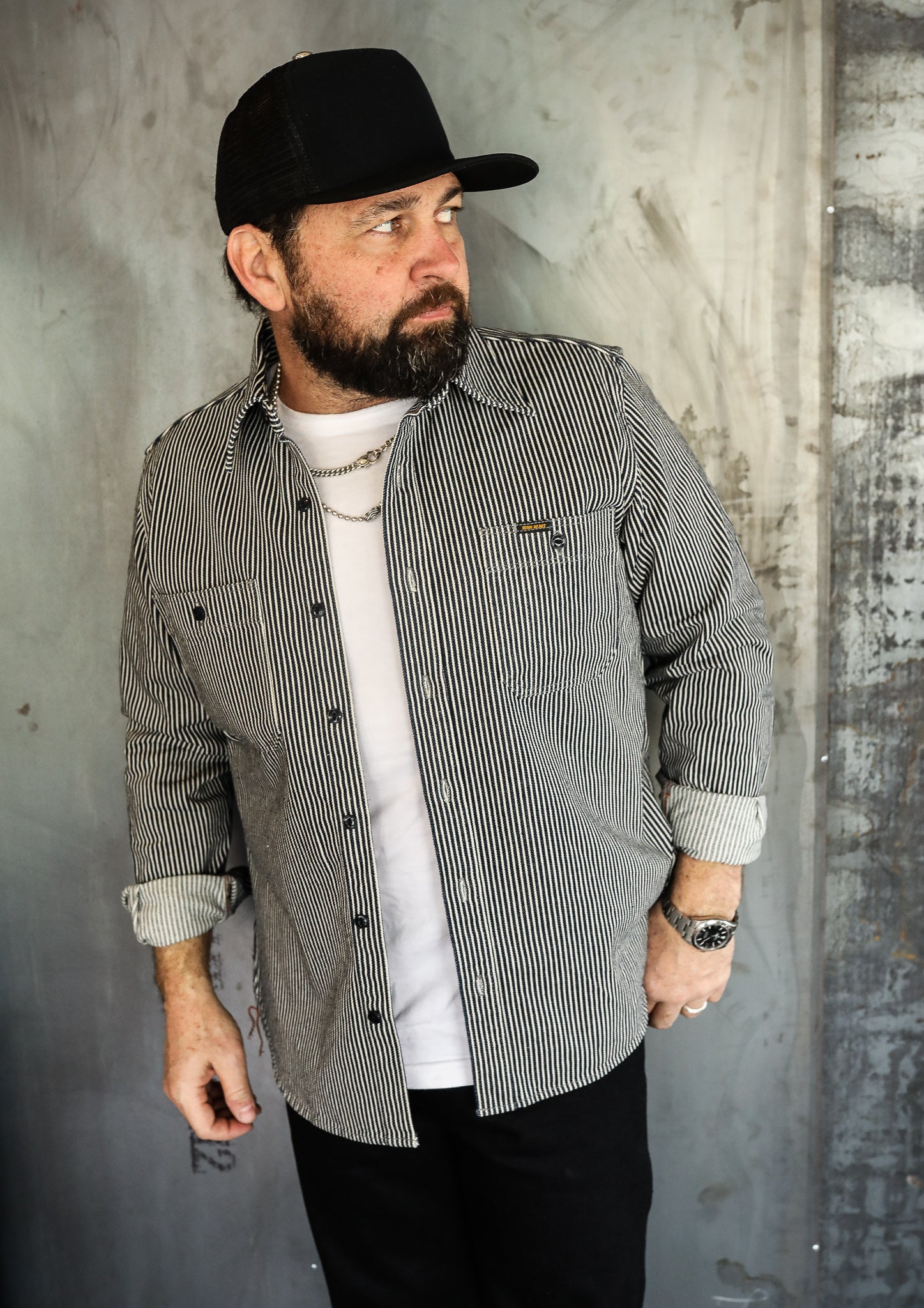 Man wearing black and white striped work shirt, white tee, black cap, and silver watch against industrial wall