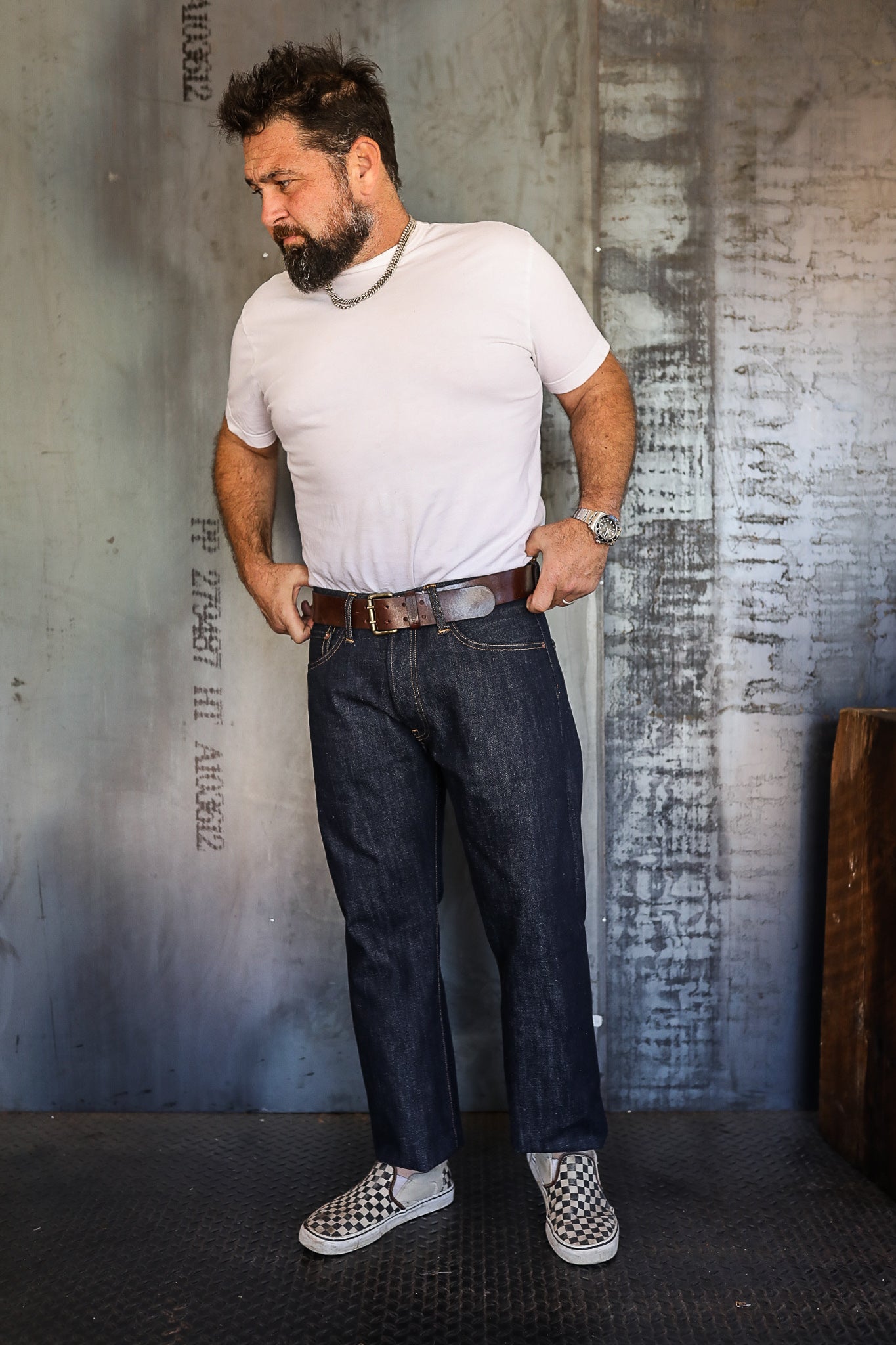 Man wearing white t-shirt, dark denim jeans, brown leather belt, silver chain necklace, and checkered slip-on shoes in industrial setting