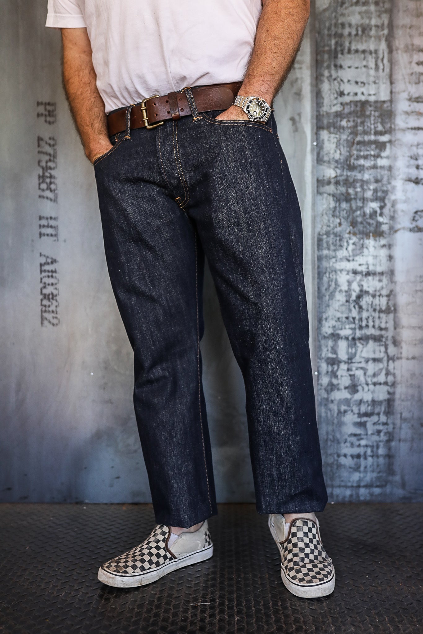Man wearing dark raw denim jeans with brown leather belt, white t-shirt, checkerboard slip-on sneakers, and silver wristwatch